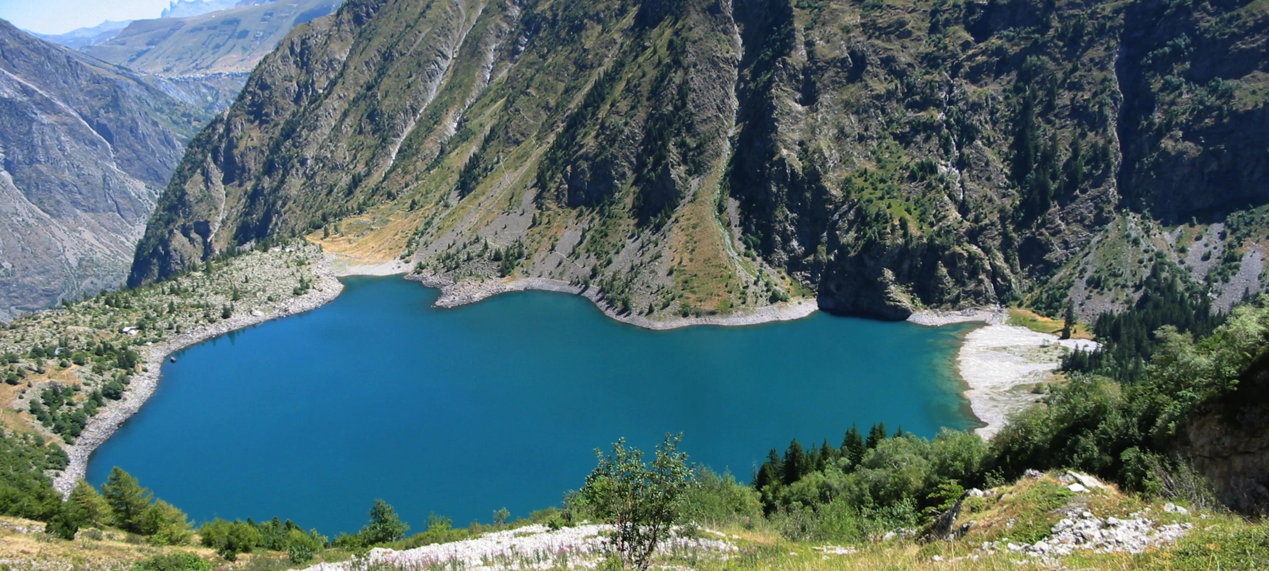 A large turquoise lake surrounded by steep mountain slopes and rocky terrain with green vegetation in the foreground.
