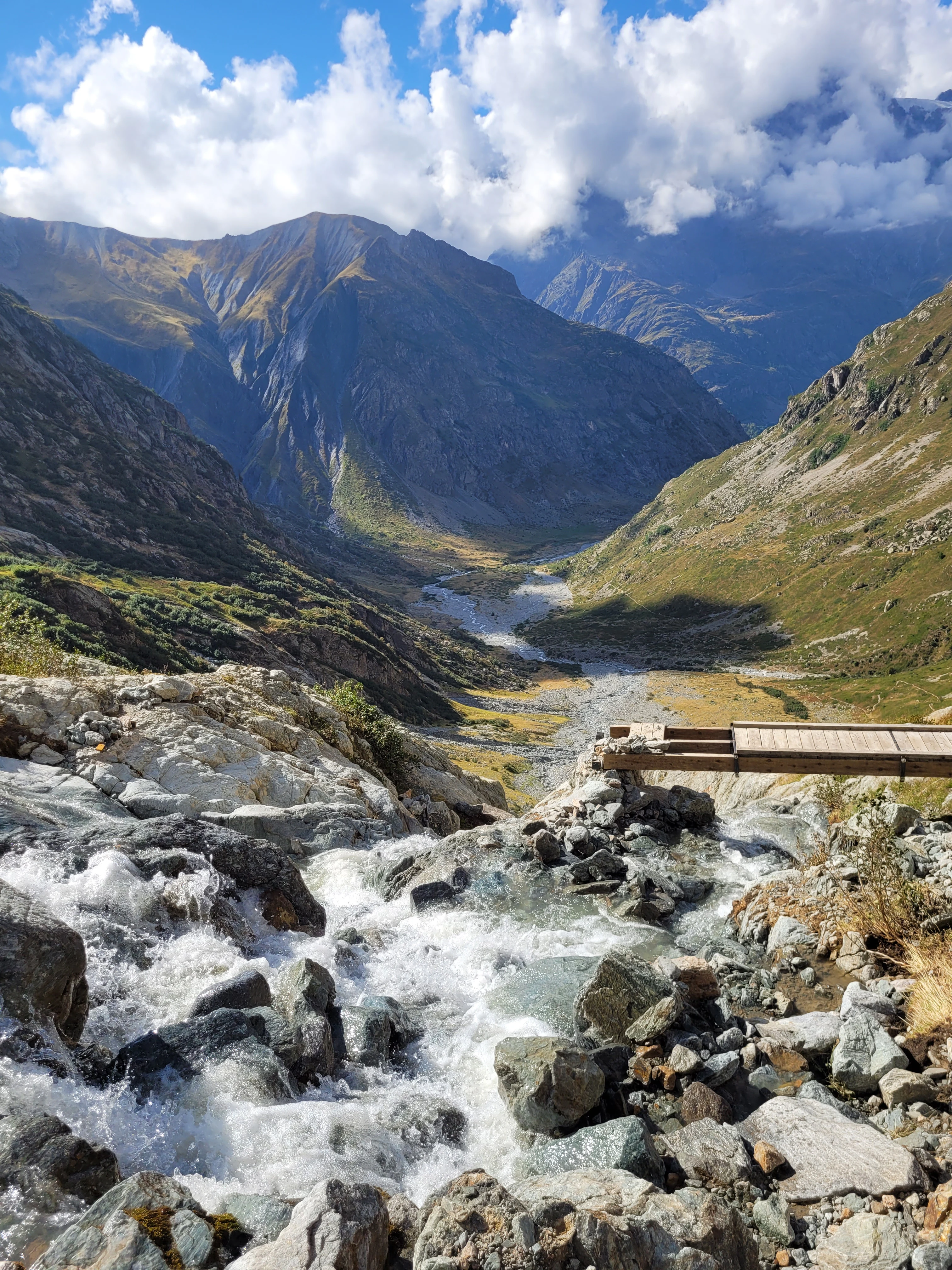 Mountain valley with a river flowing over rocks, a wooden footbridge, and mountain peaks in the background.