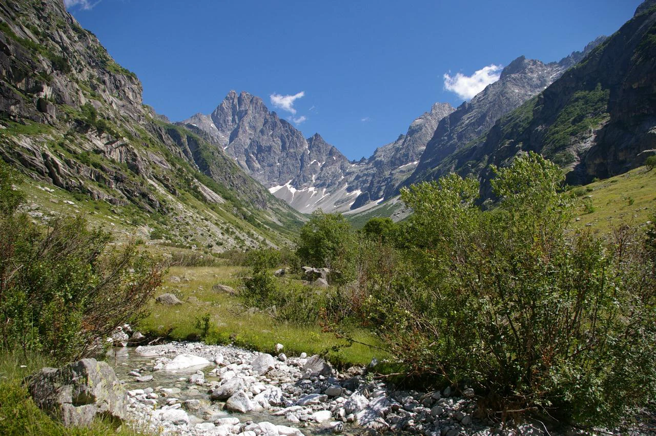 A mountain valley with a stream flowing through rocky terrain, surrounded by green slopes and rocky peaks under a clear blue sky.