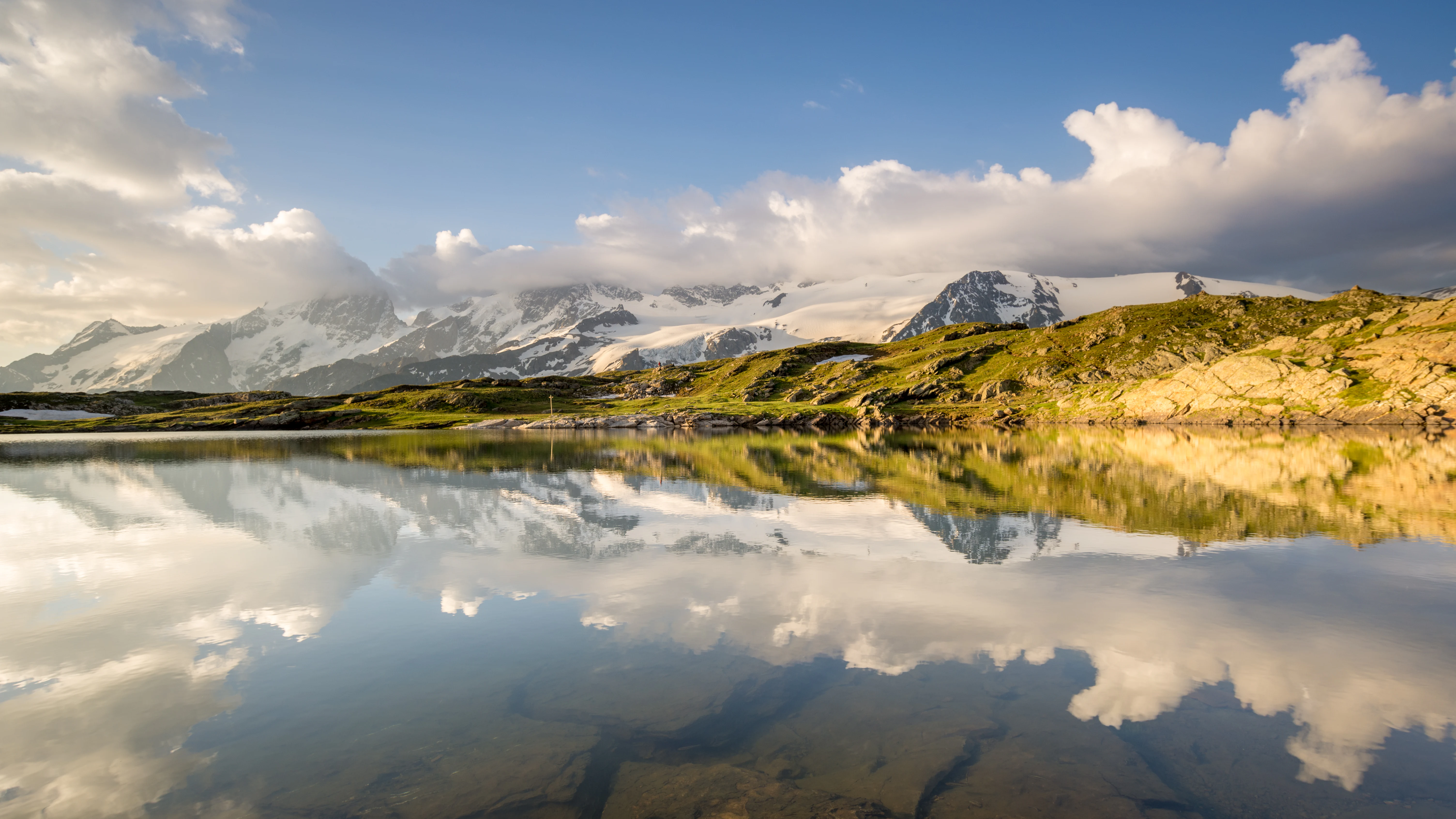 A calm lake reflecting snow-covered mountains and clouds under a partly cloudy sky