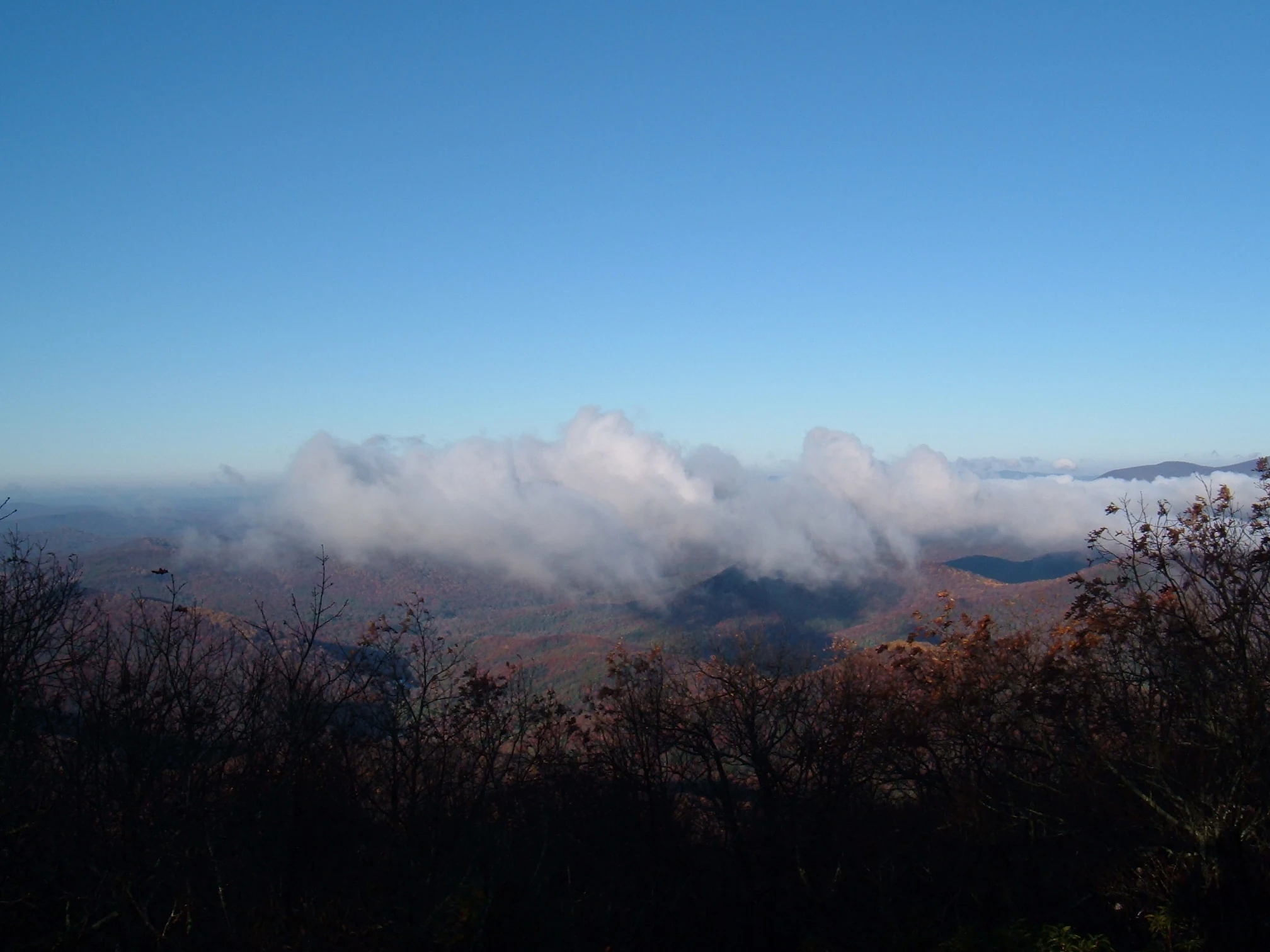 A wide view from the top of a mountain, looking out over rolling hills partially obscured by low-hanging clouds under a clear blue sky, with autumn-colored trees in the foreground.