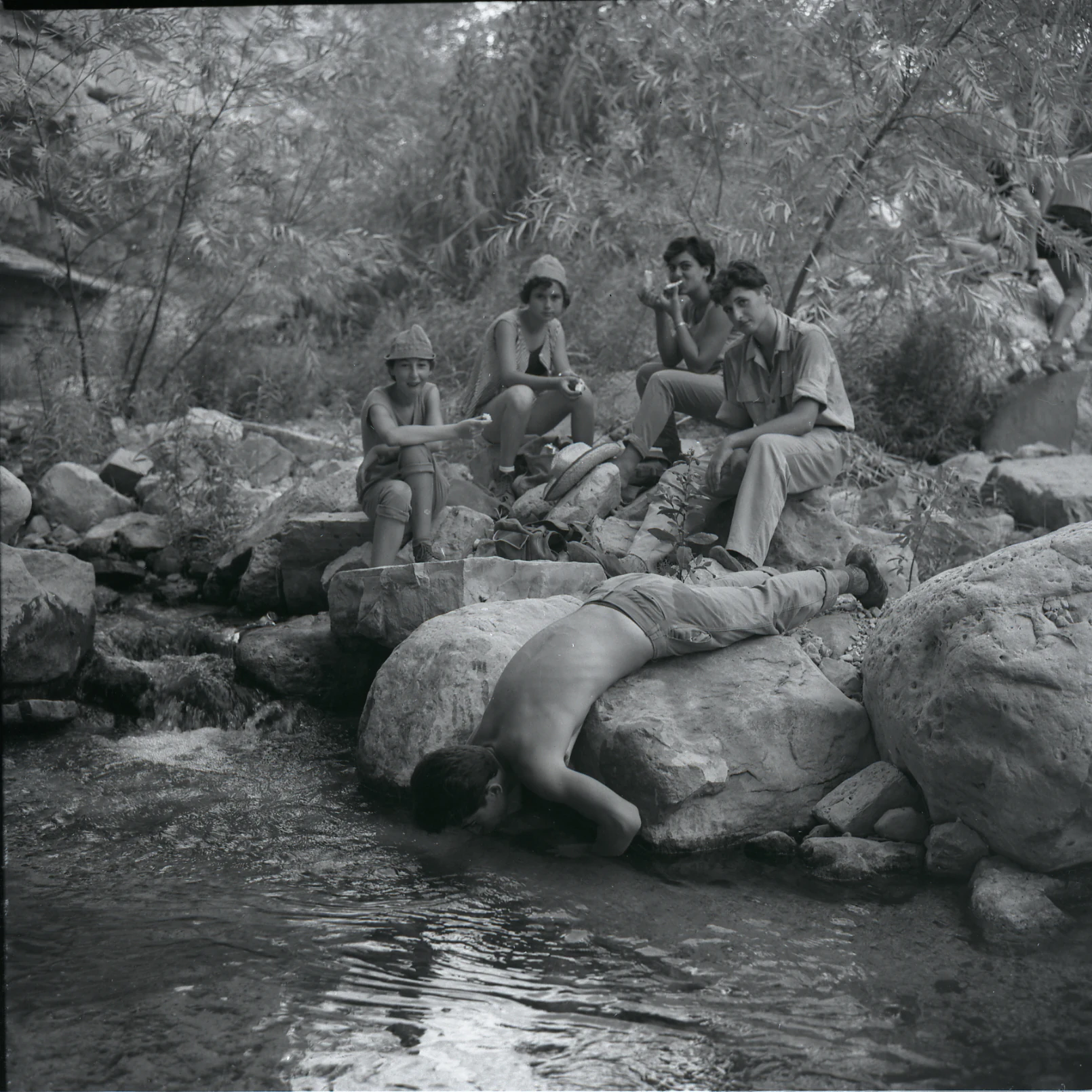 Black-and-white photograph showing several people near a stream in a rocky, vegetated area with vegetation in the background
