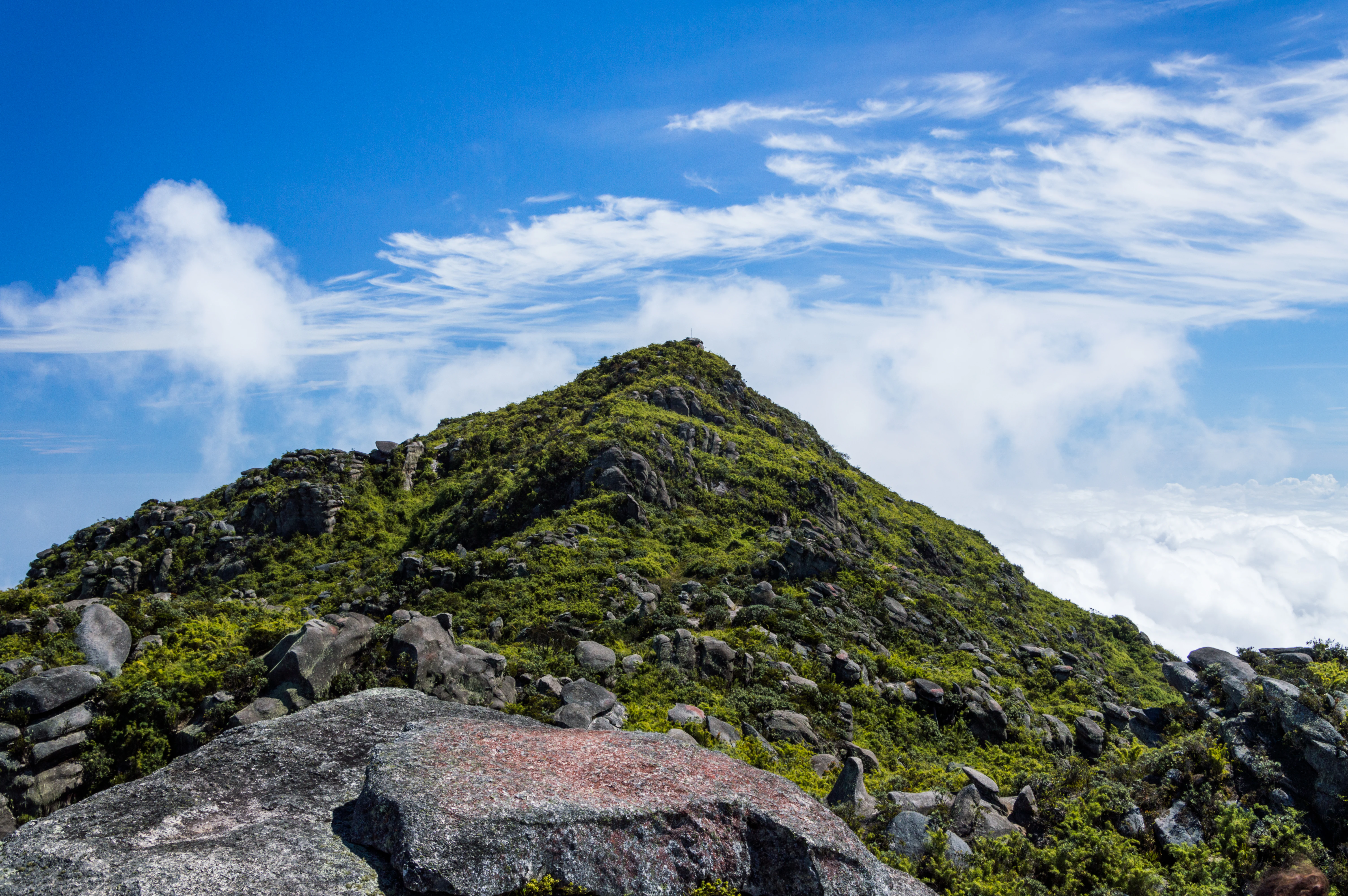 Rocky mountain peak covered in green vegetation under a blue sky with scattered clouds.