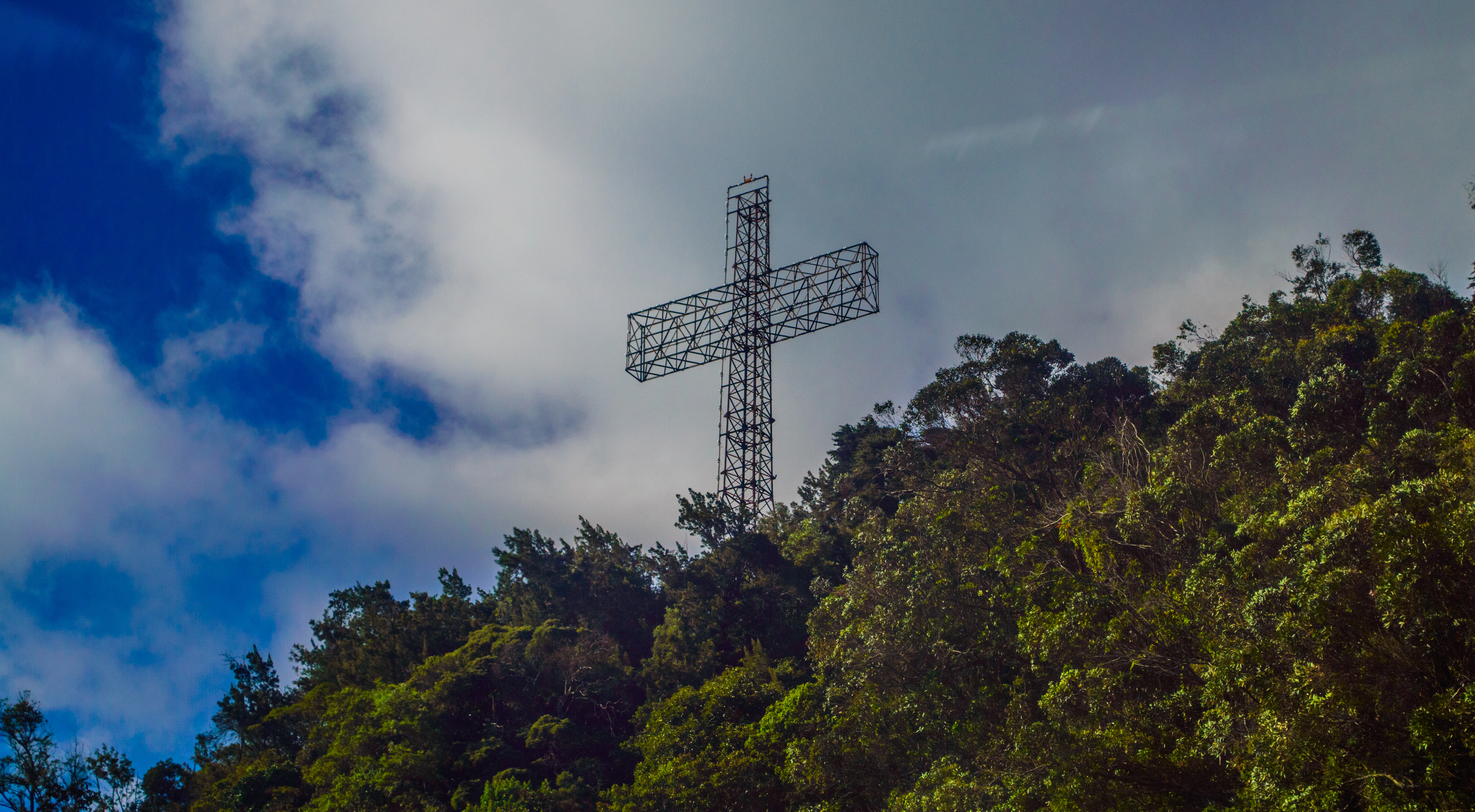 Tall metal cross structure atop a forested hill against a partly cloudy sky