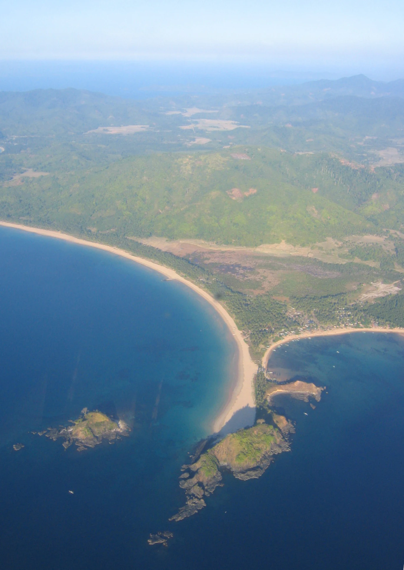 Aerial view of a thin sandy isthmus separating two bodies of blue water, with a lush green hilly peninsula extending into the ocean.
