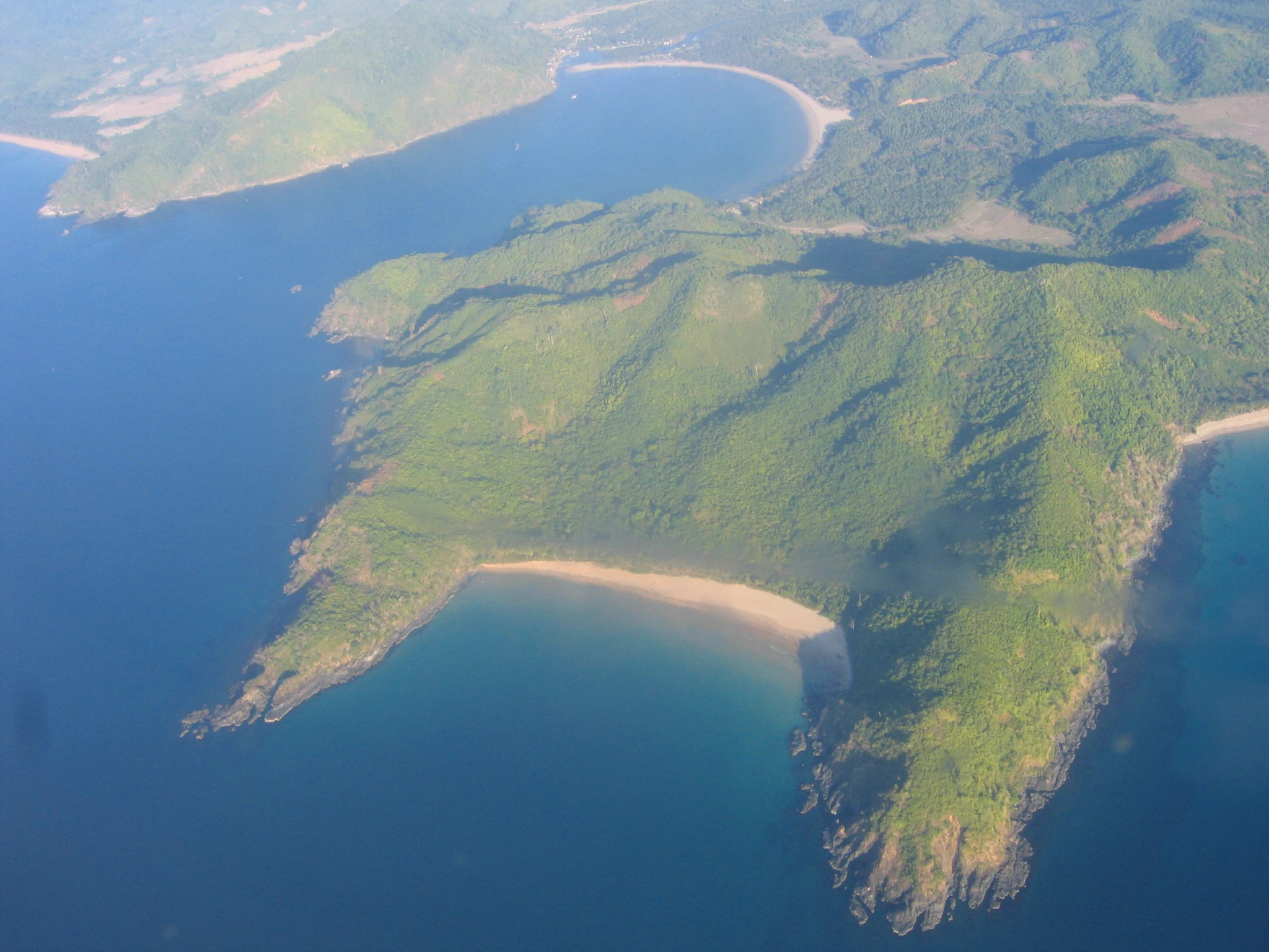 An aerial view showing a lush green peninsula extending into blue ocean water, with sandy beach coves and surrounding mountainous terrain.