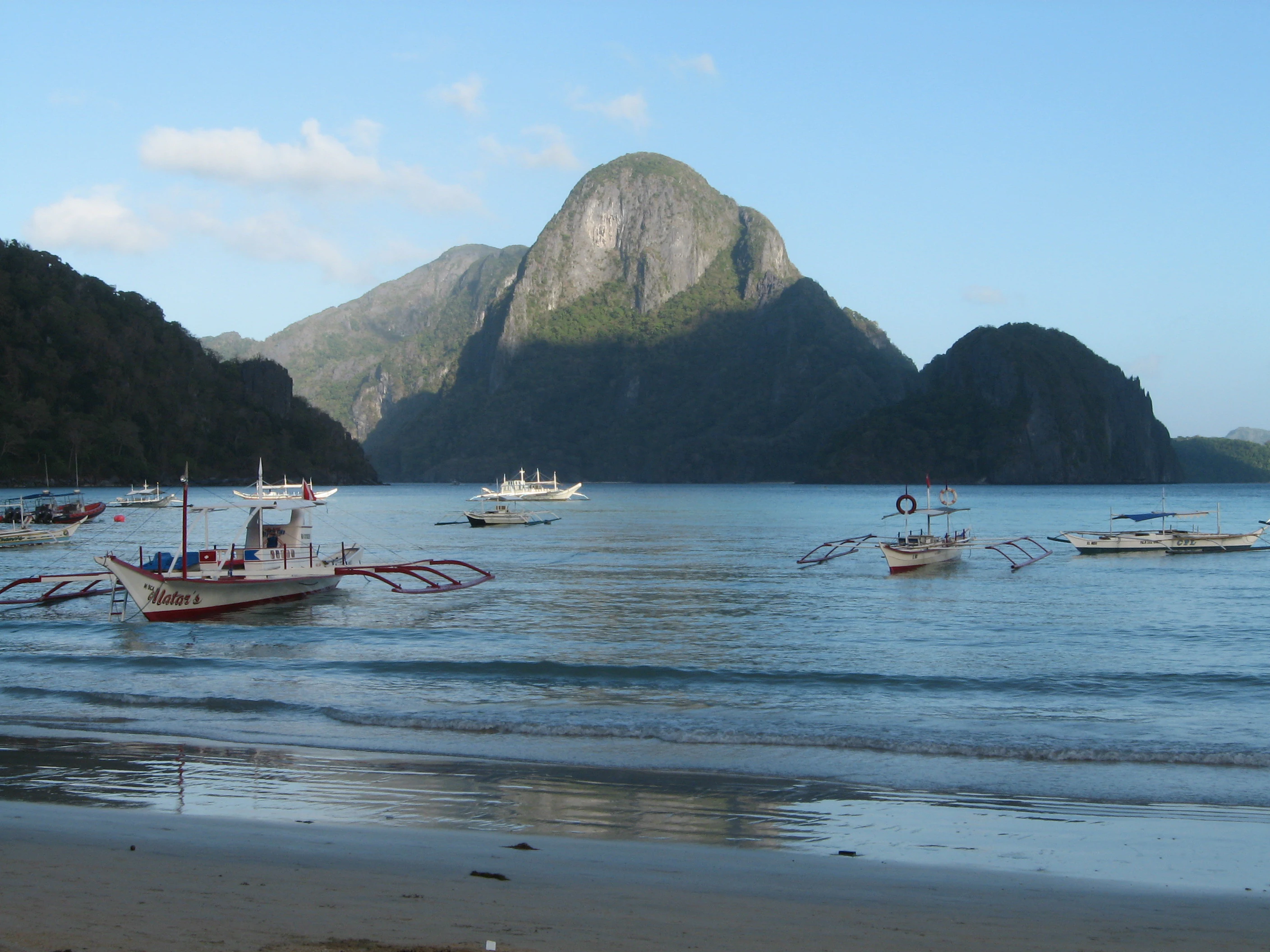 Several traditional Filipino outrigger boats float on calm water in front of large limestone cliffs under a clear blue sky.