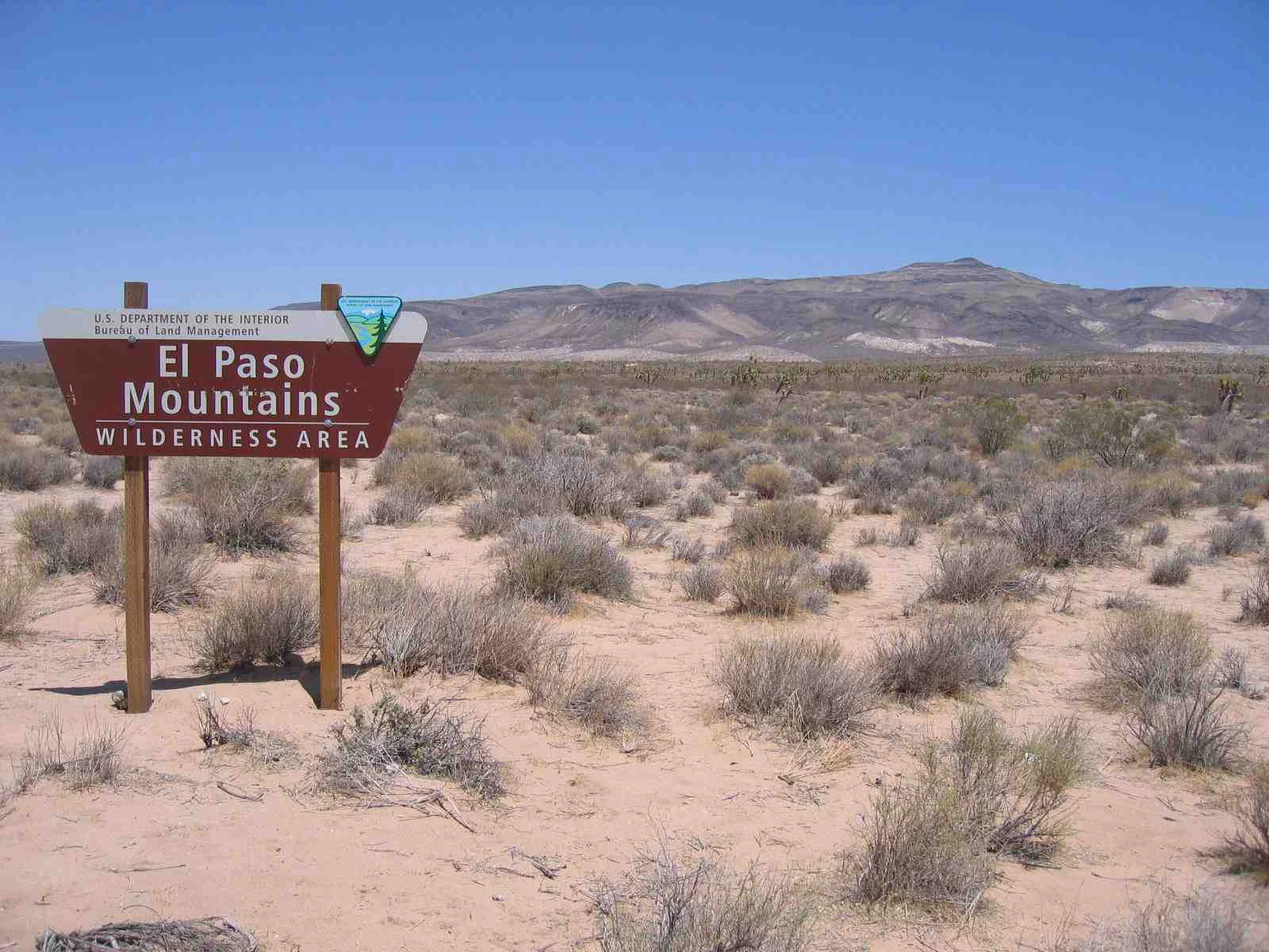 A brown Bureau of Land Management sign reading El Paso Mountains Wilderness Area stands in a sandy, shrub-filled desert landscape under a clear blue sky.