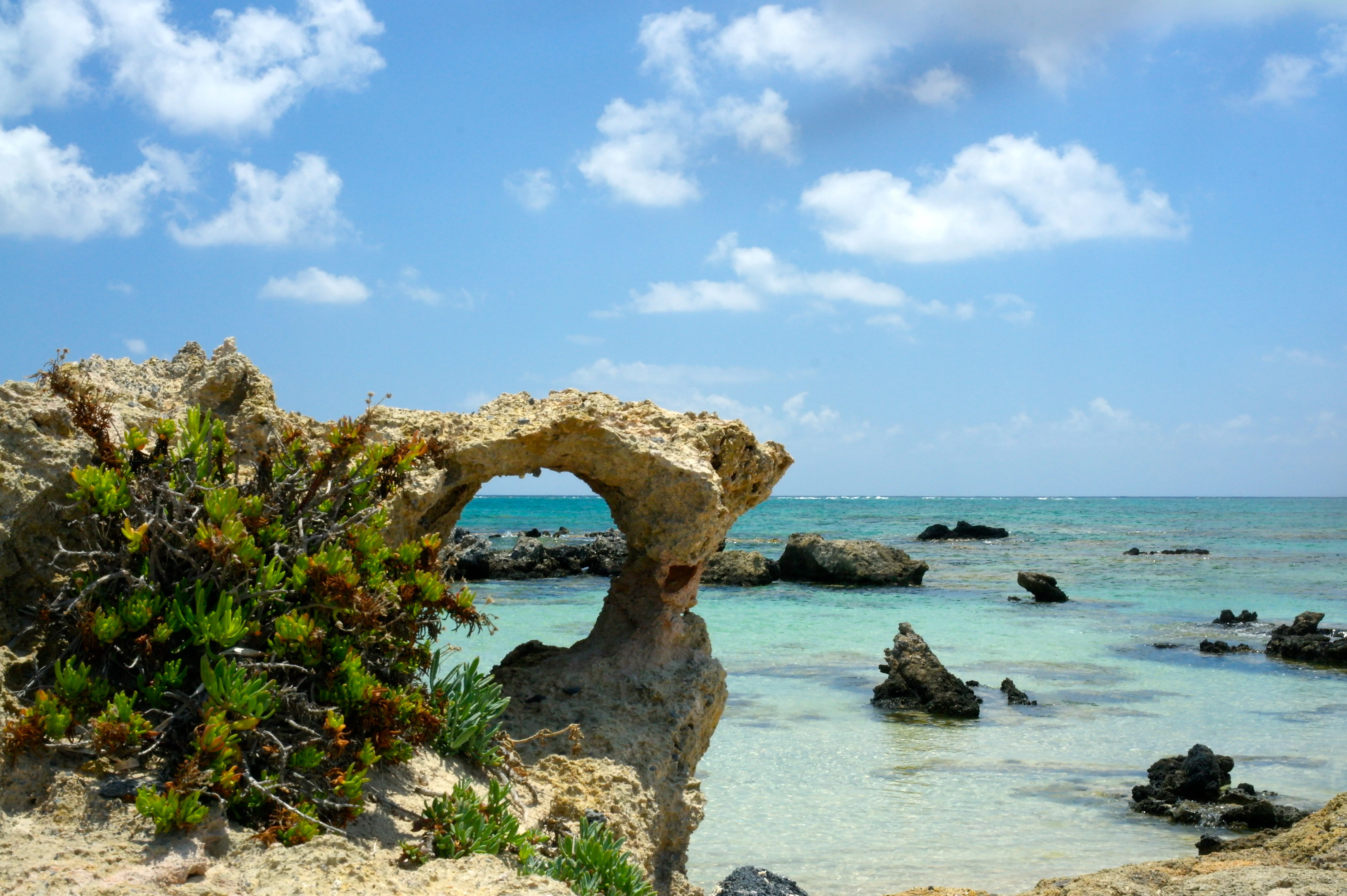 A weathered rock arch stands on a rocky coastline overlooking shallow turquoise sea water under a blue sky with scattered clouds.