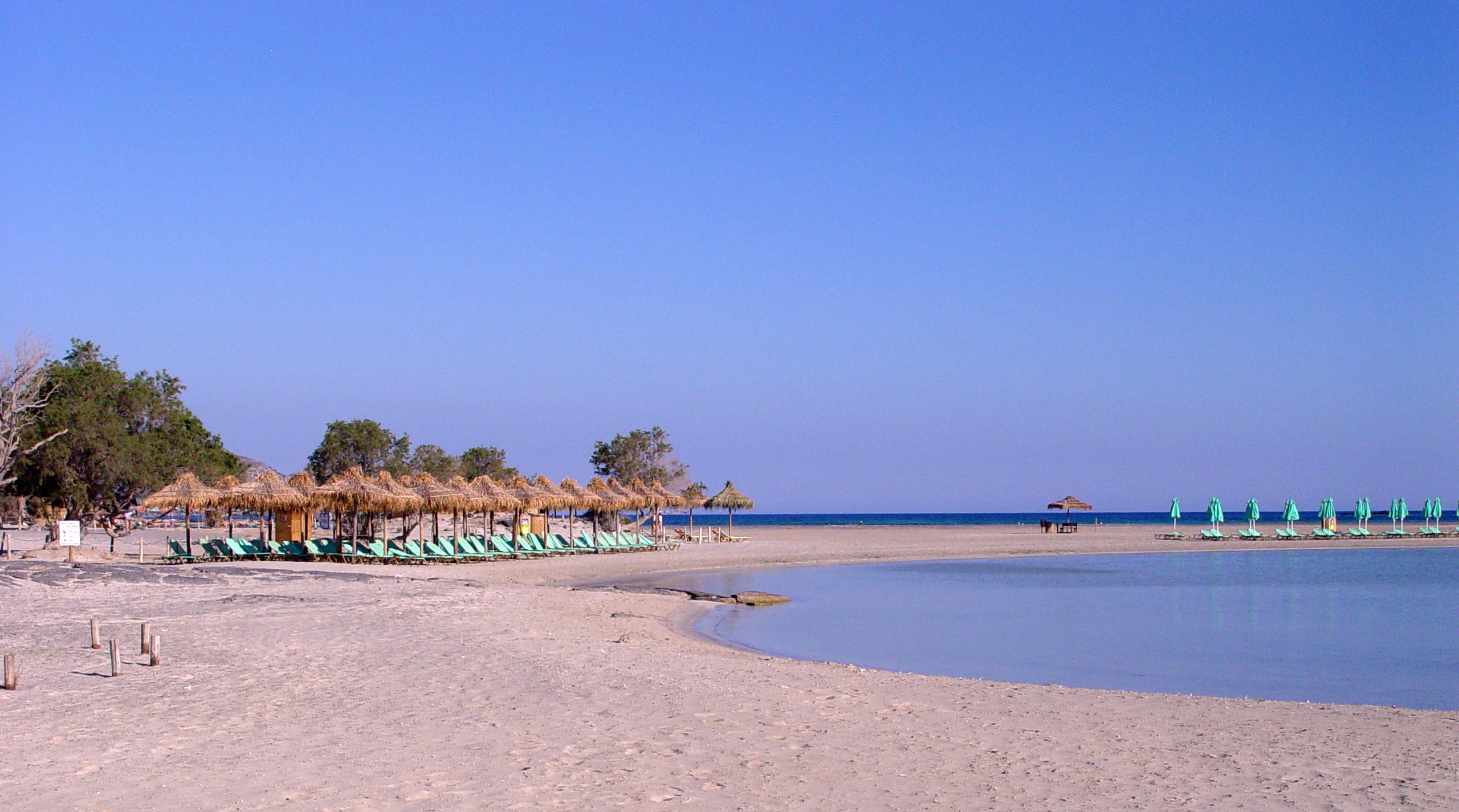 A wide-angle, eye-level shot of a sandy beach with rows of thatch umbrellas and lounge chairs, bordering a calm, shallow turquoise-blue lagoon under a clear, bright blue sky.