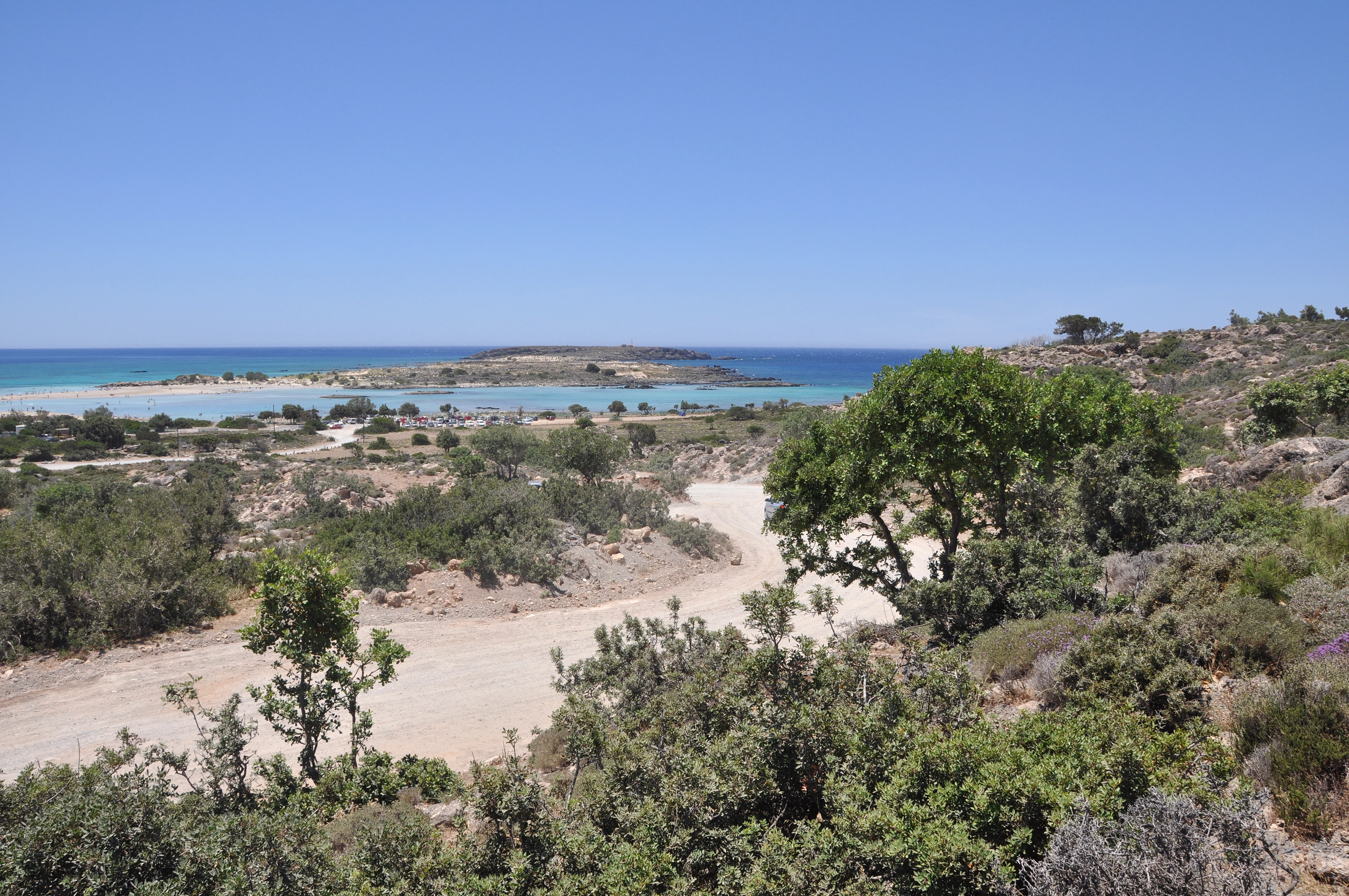 A sunny coastal landscape in Crete showing a dirt path leading toward a sandy beach, shallow turquoise water, and a small island in the distance under a clear blue sky.