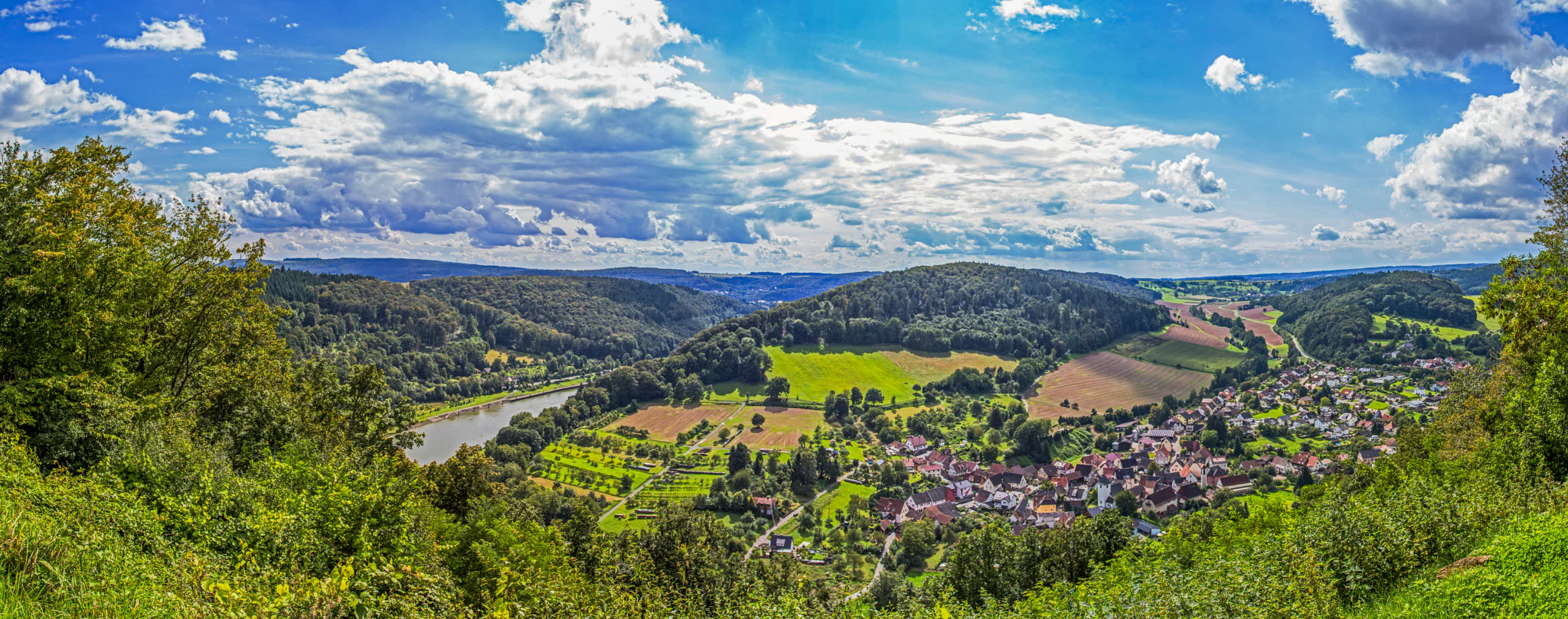 A wide, panoramic view from a high vantage point overlooking the town of Neckargerach, nestled in the green valley of the Neckar River, surrounded by forested hills under a blue sky with scattered white clouds.