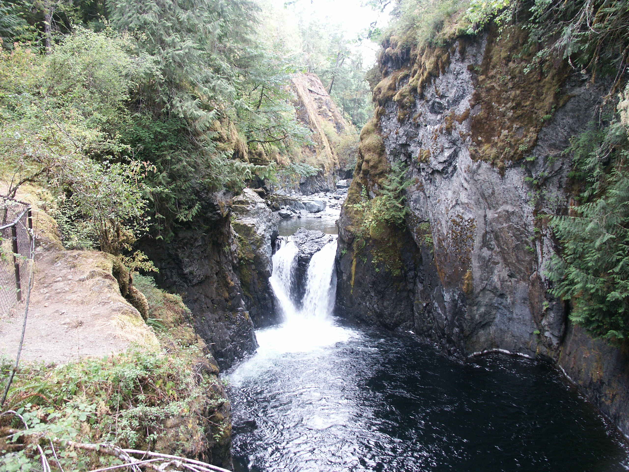 A view of a waterfall flowing through a narrow, mossy rock canyon, surrounded by dense forest foliage, with a hiking trail railing visible on the left.