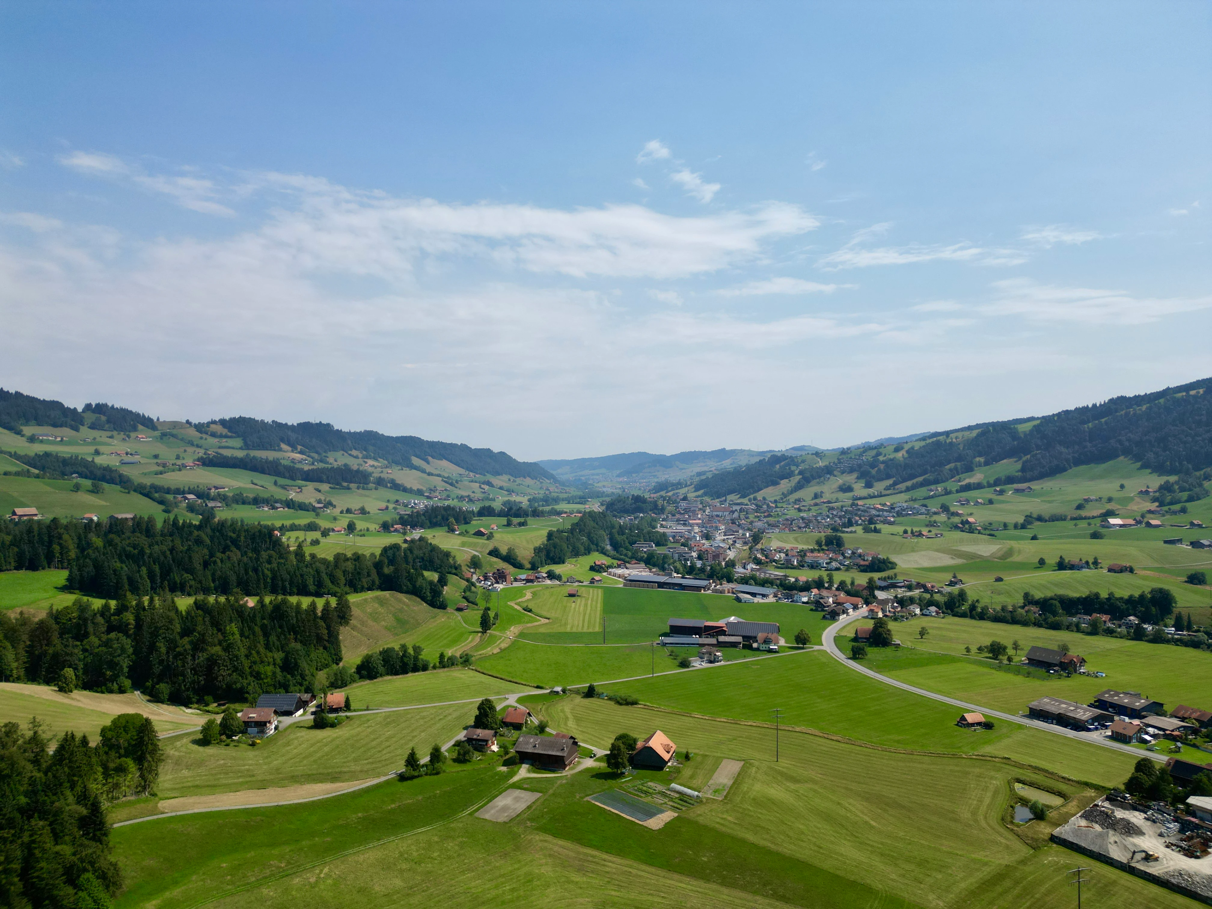 A high-angle, wide-angle view of a Swiss valley featuring rolling green meadows, patches of coniferous forest, and a small town clustered in the distance under a blue sky.