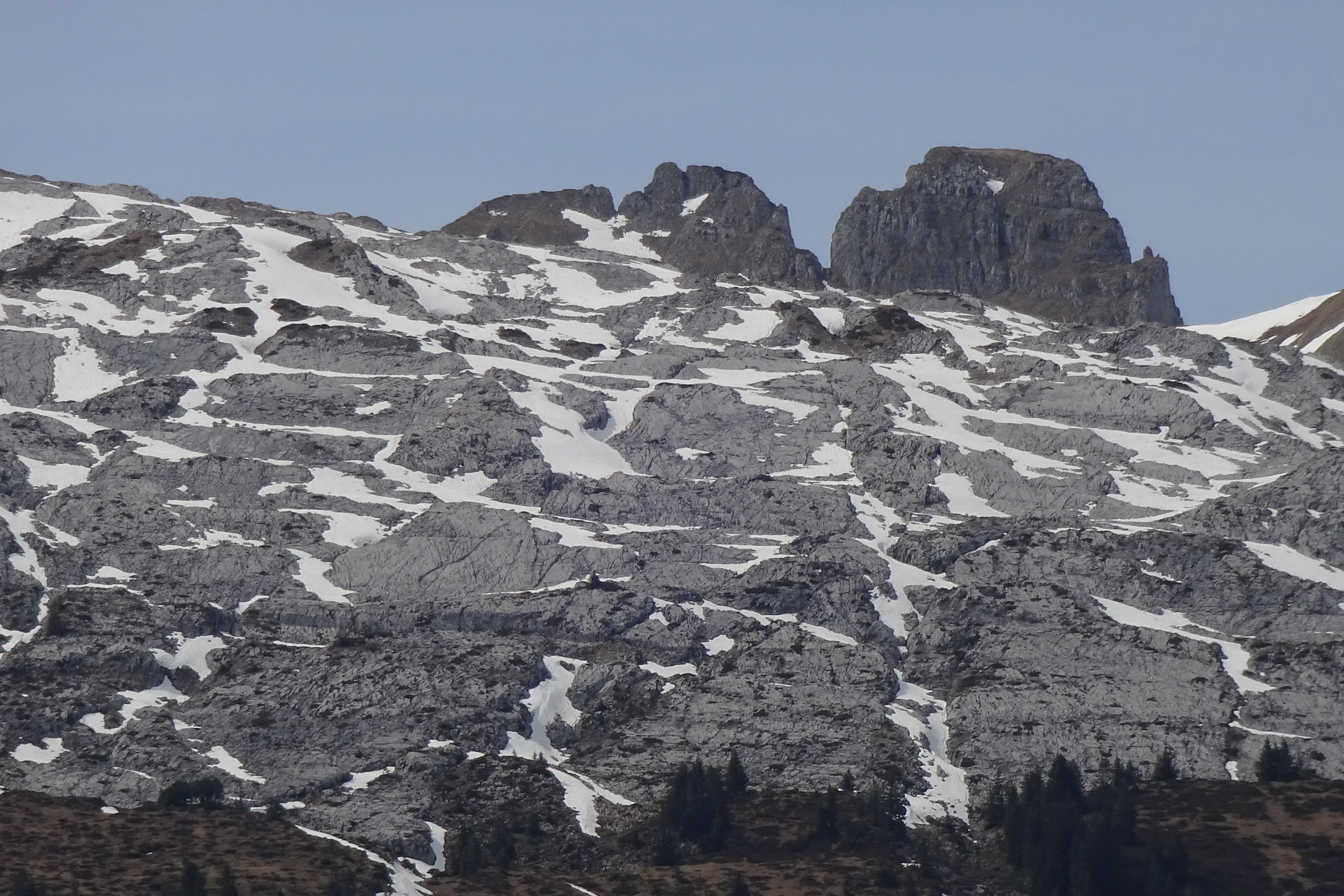A wide mountain slope covered in grey limestone rock and scattered patches of snow, with prominent craggy peaks rising against a clear sky.