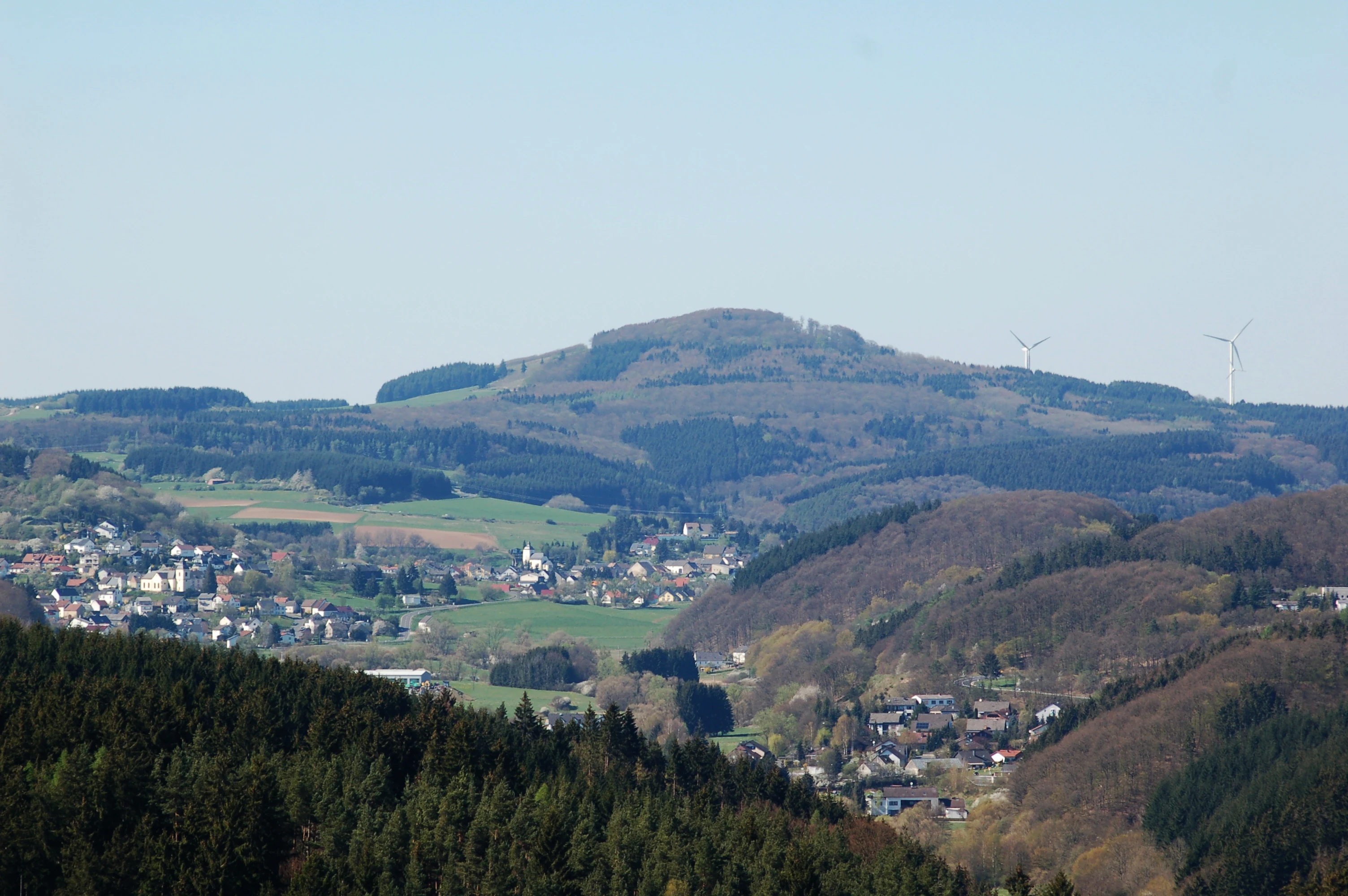 A wide shot of a rolling, forested volcanic hill with small towns and green fields in the valley below under a clear sky.