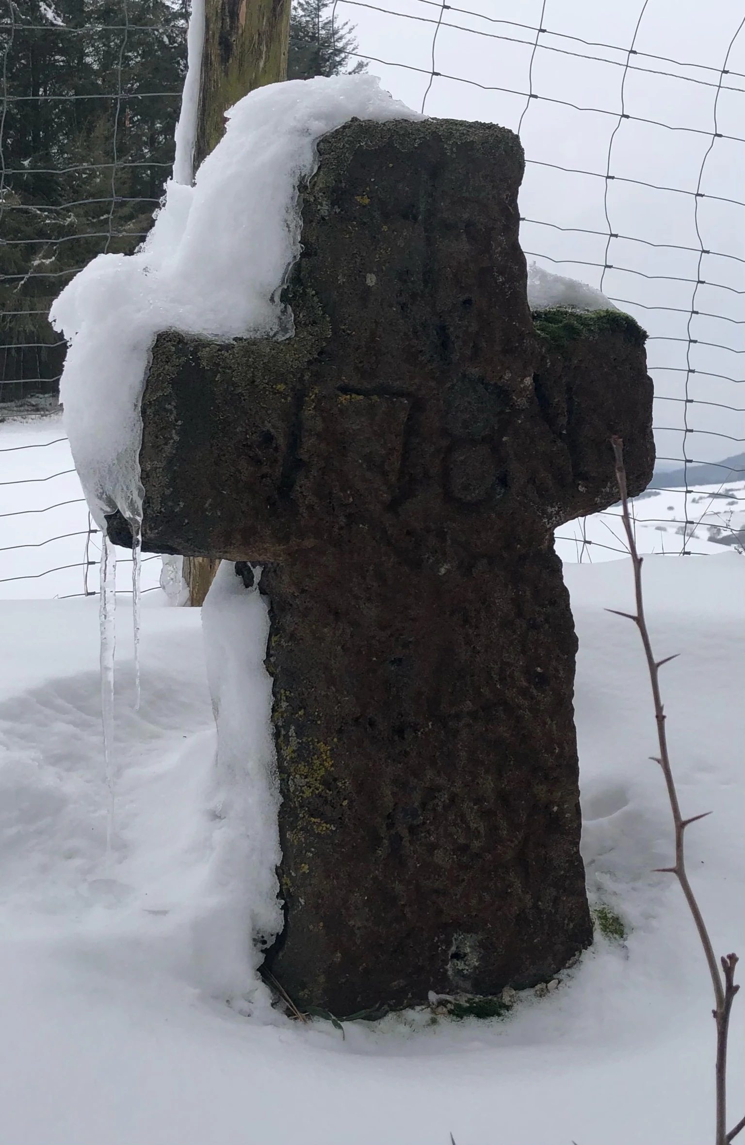 A weathered dark stone cross with faint engraved numbers standing in a snow-covered field with an icicle hanging from the top left corner, set against a wire fence background.