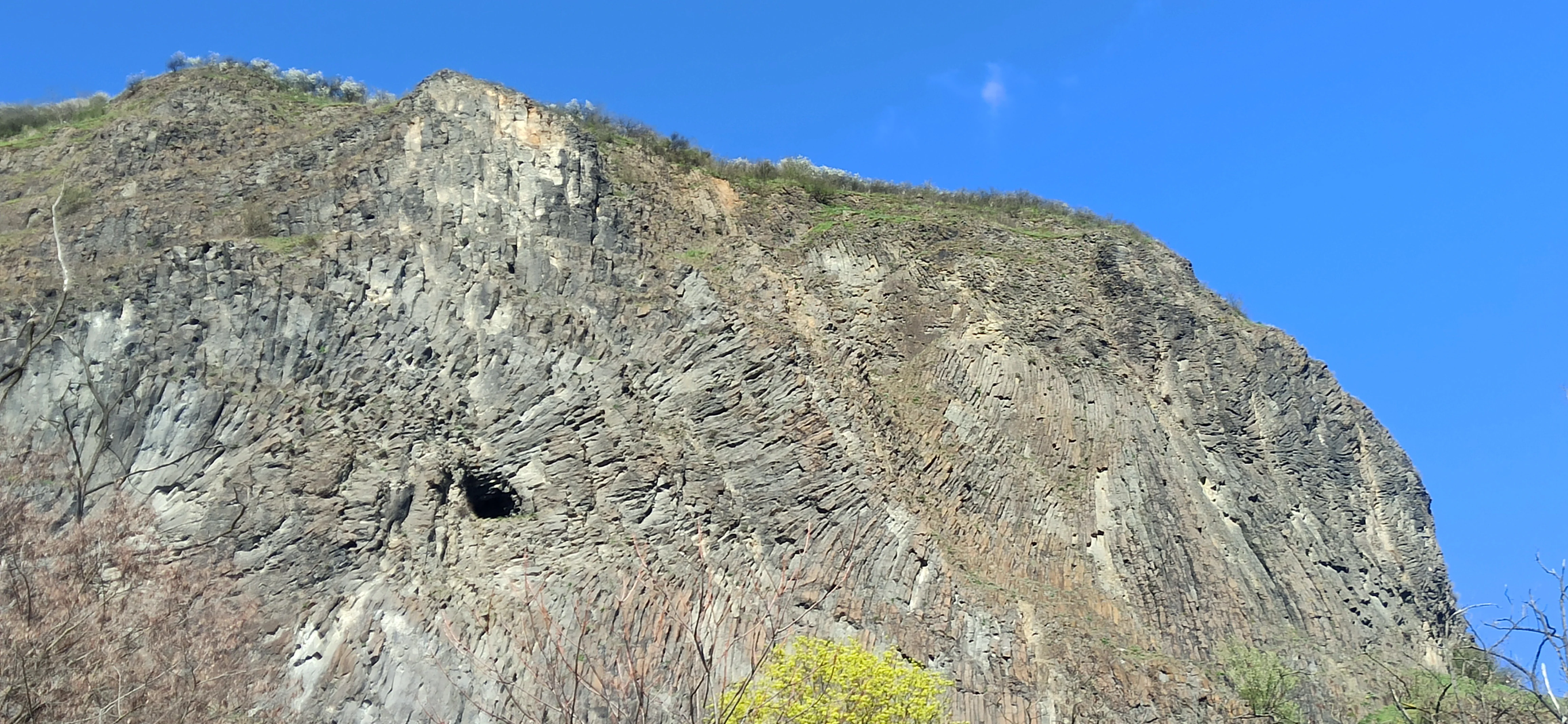 A steep, craggy cliff face showing distinct columnar basalt rock formations against a clear blue sky.