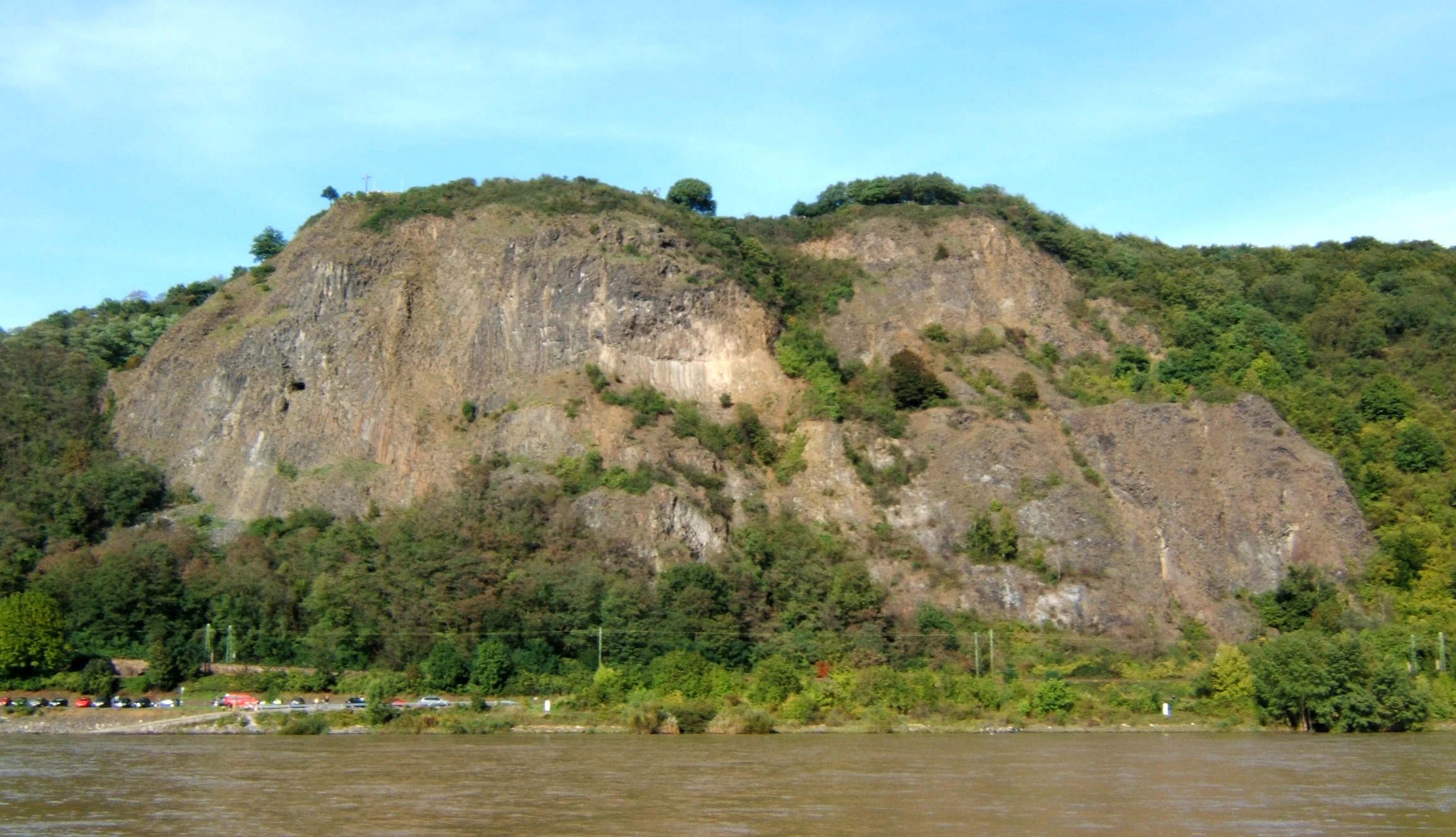 A wide view of the Erpeler Ley basalt rock formation rising prominently above the Rhine River, with trees and greenery covering parts of the cliff face.