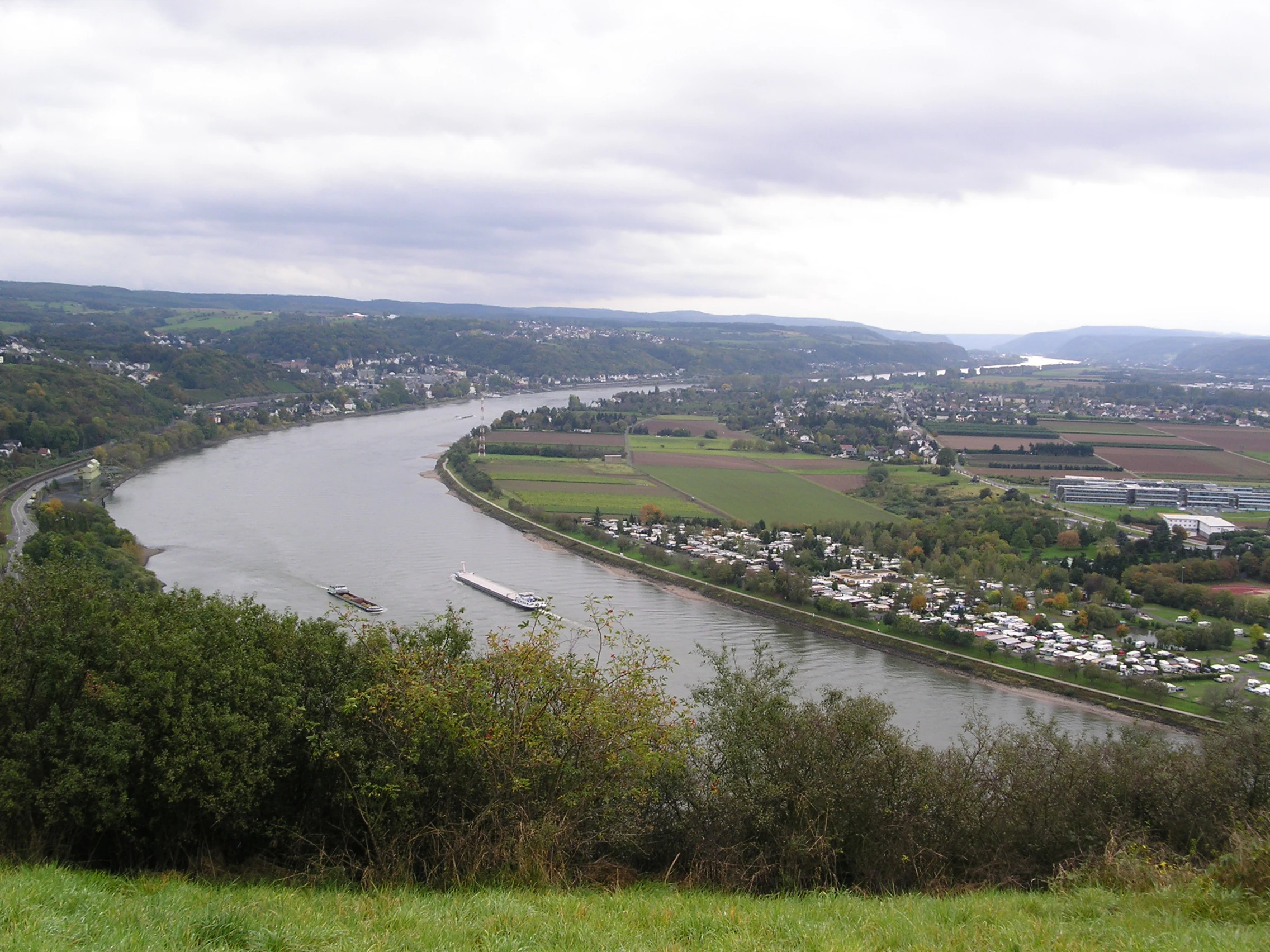 An elevated panoramic view looking down over a winding river with a barge, surrounded by green fields, small settlements, and dense vegetation under a cloudy sky.