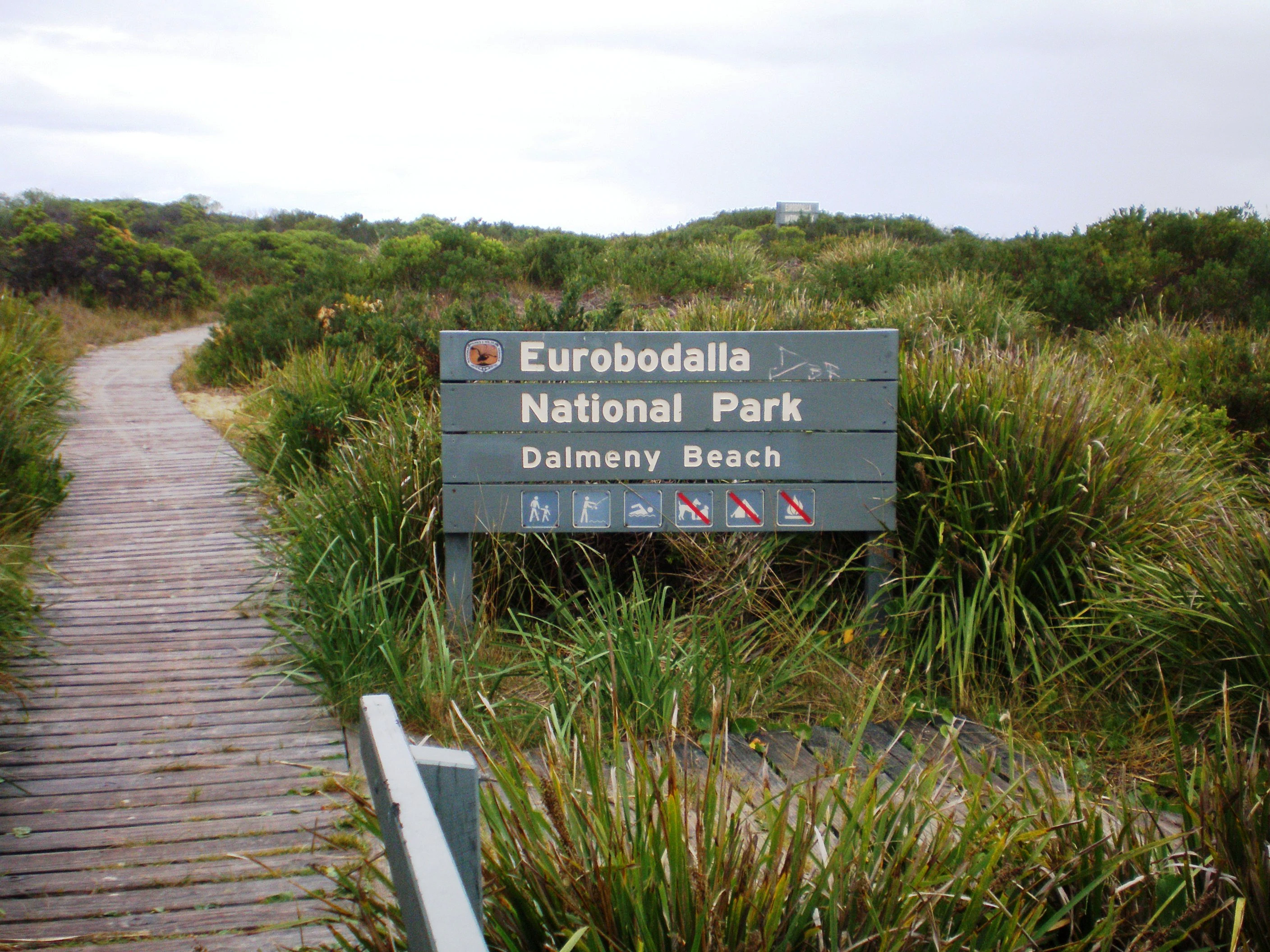 Wooden boardwalk leading to Eurobodalla National Park entrance sign, surrounded by tall grasses and coastal vegetation