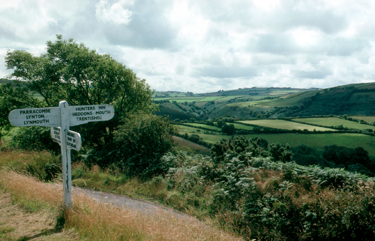 White signpost with directional signs for Princetown, Lynton, and other locations, set against a backdrop of green rolling hills and fields