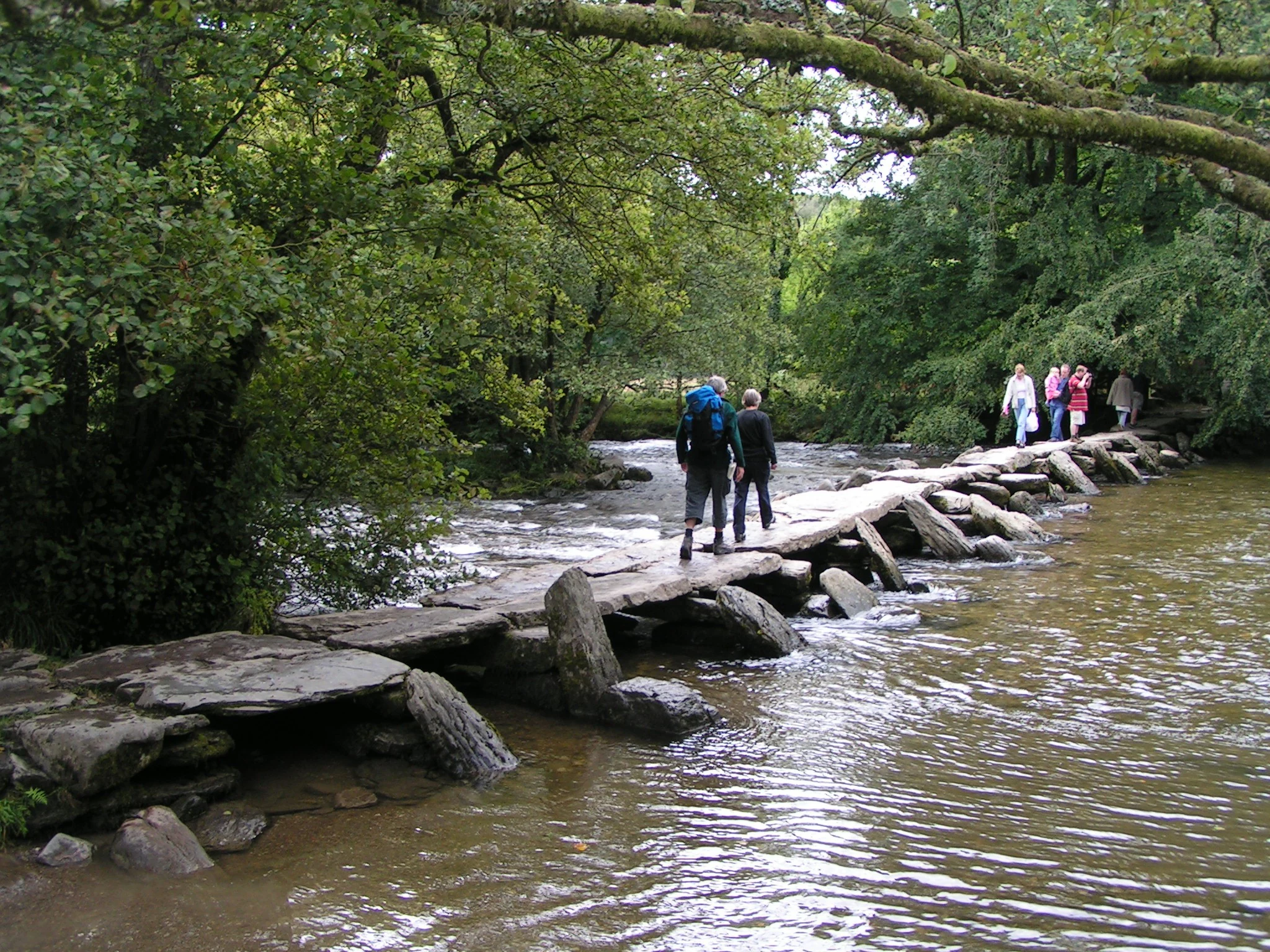 Stone clapper bridge over a river with people walking on it, surrounded by trees