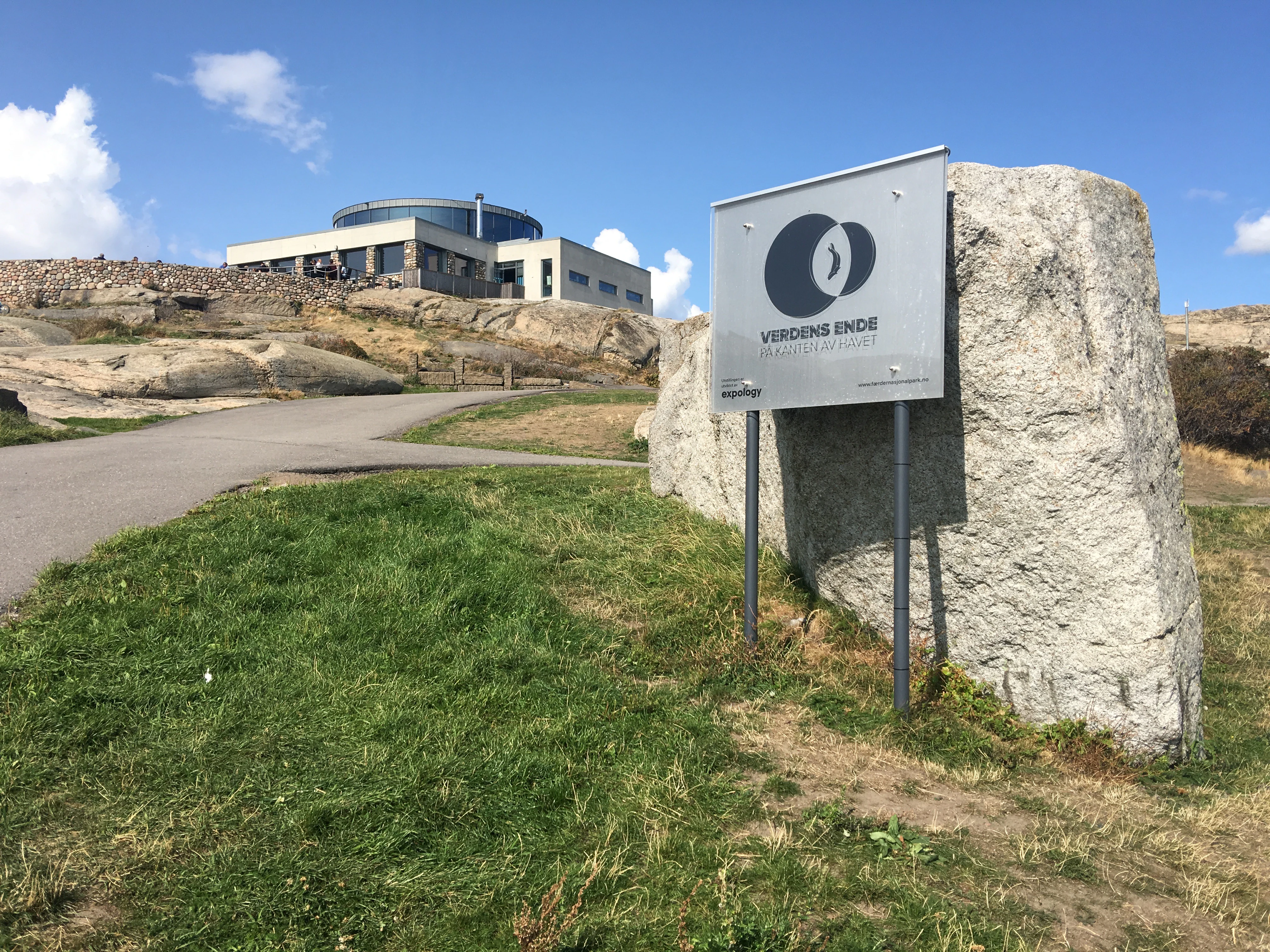 A metal sign for Færder National Park visitor centre mounted on a stone structure, with a building and rocky landscape in the background under a clear blue sky