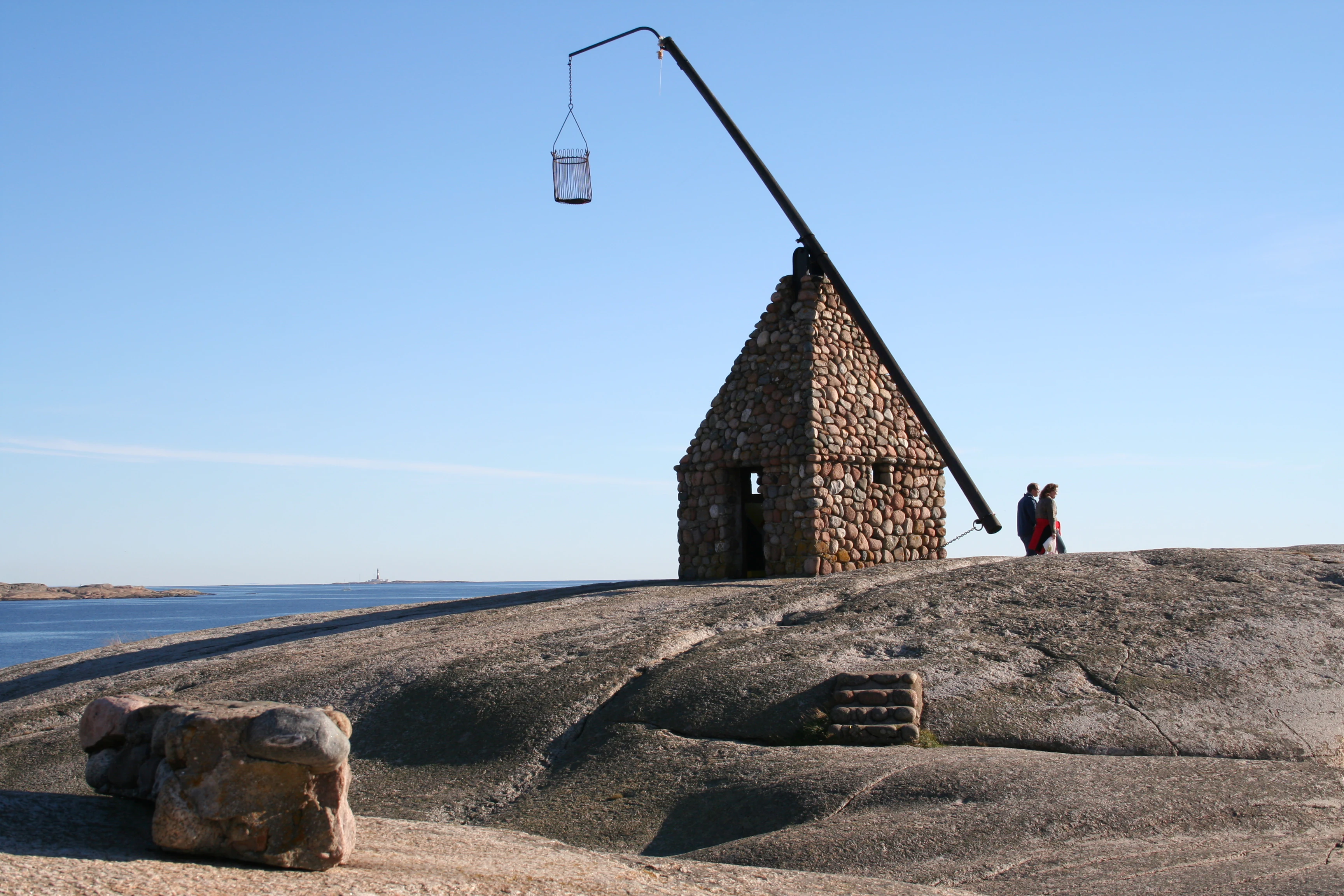 Stone building with a long arm holding a basket, two people standing nearby, on a rocky hill overlooking water under a clear blue sky