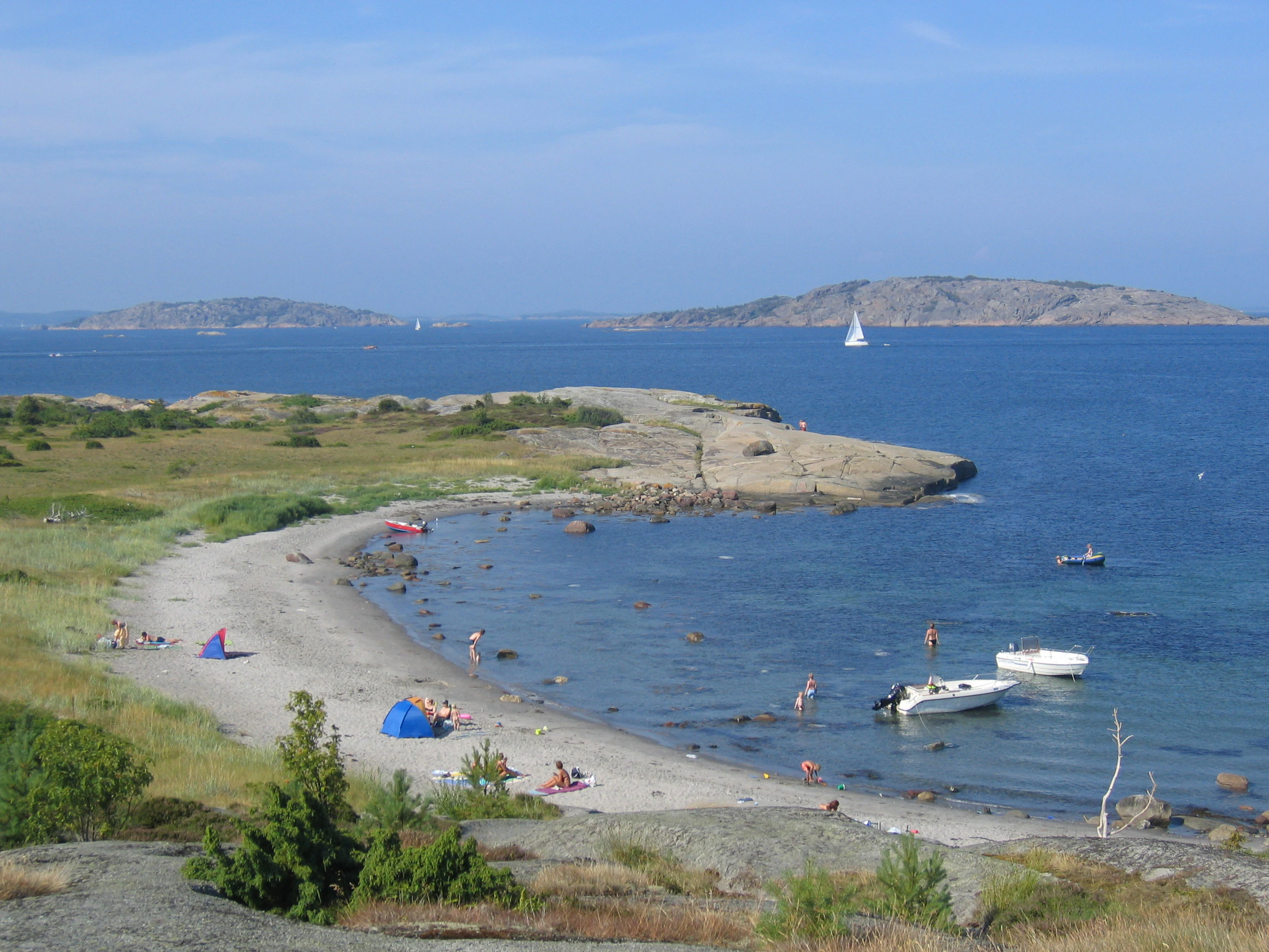 Sandy beach with people sunbathing, small boats anchored in calm water, grassy areas, and islands in the distance.
