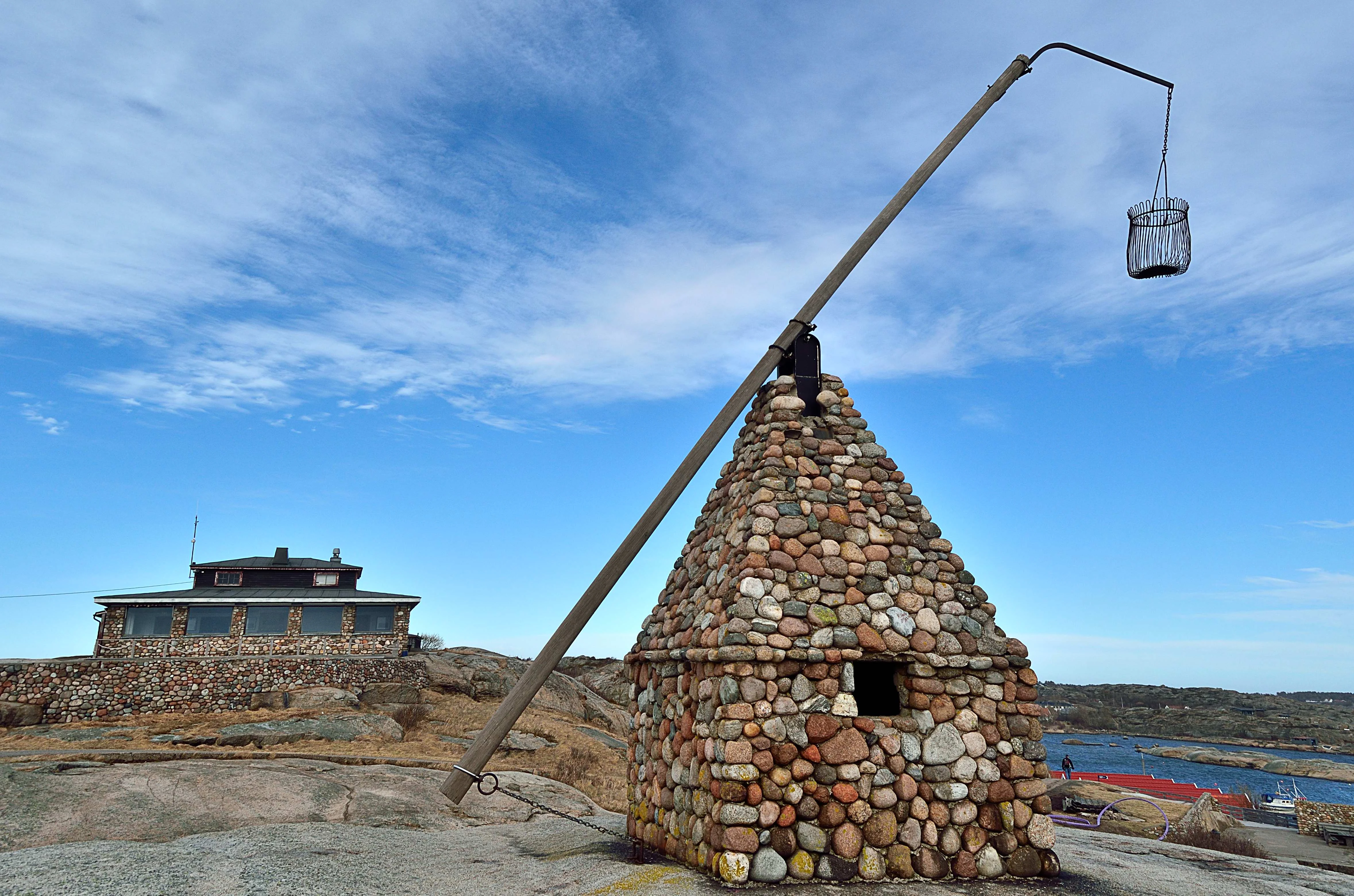 Stone tower with a wooden pole holding a metal basket, adjacent to a multi-story building by the water, under a blue sky with scattered clouds
