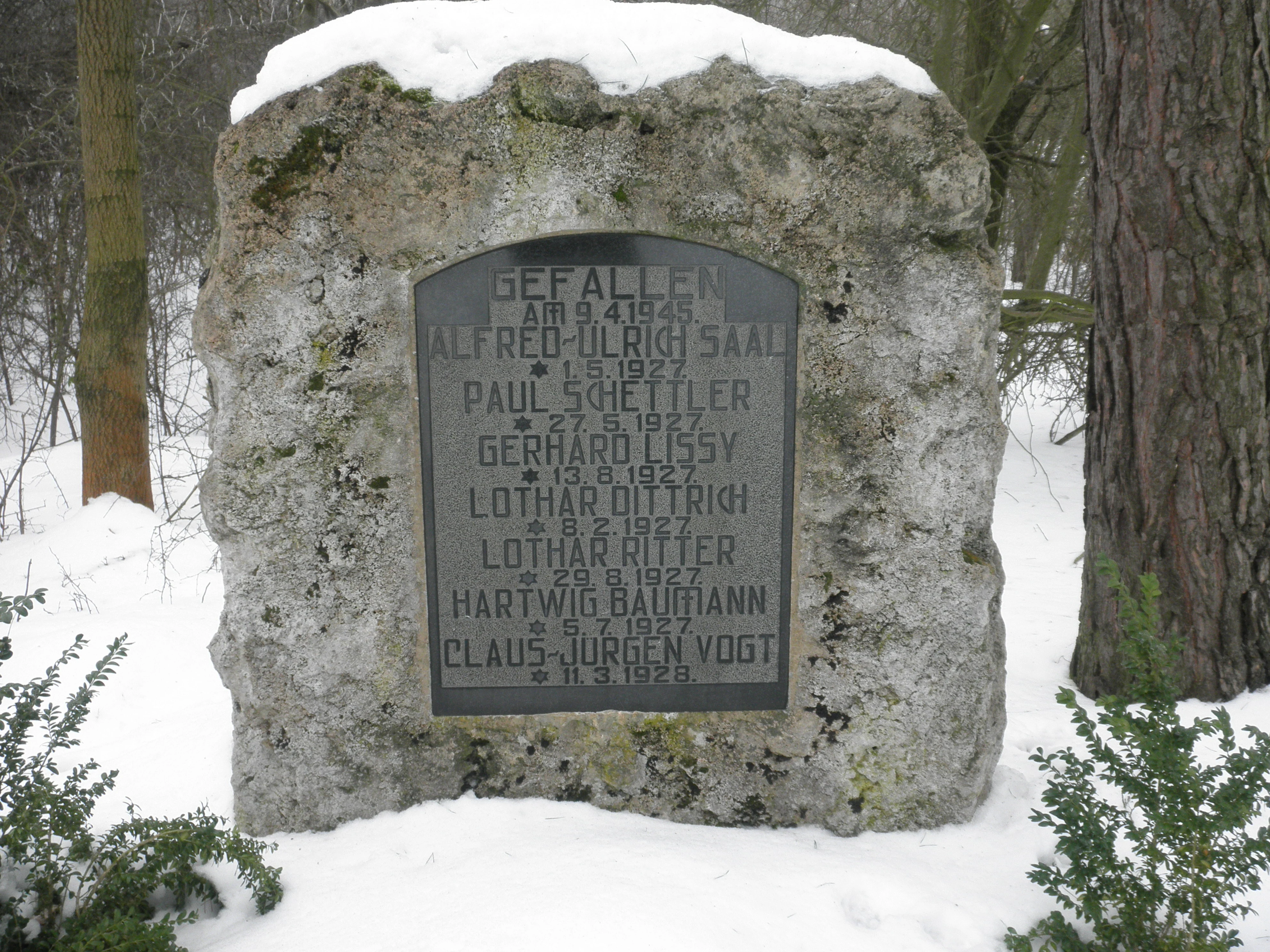A weathered stone monument with an engraved dark plaque listing names and dates, surrounded by snow in a forested area.