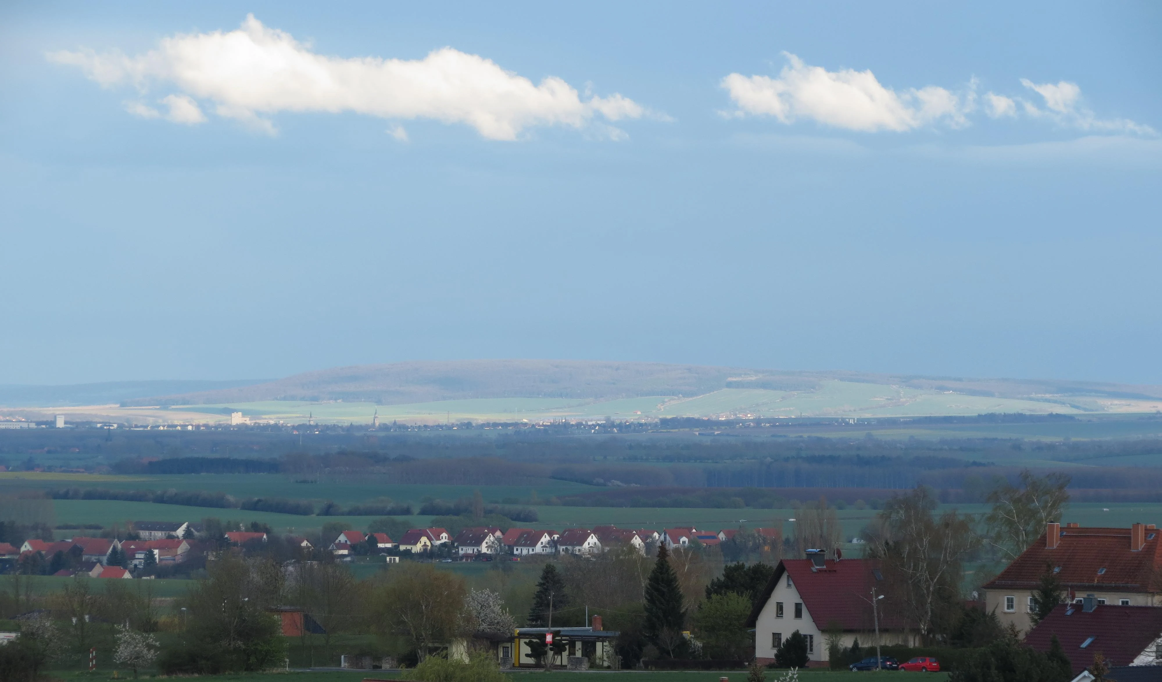 A wide, hazy landscape view of the Fahner Höhe ridge in the distance, seen across a rural area with foreground houses.