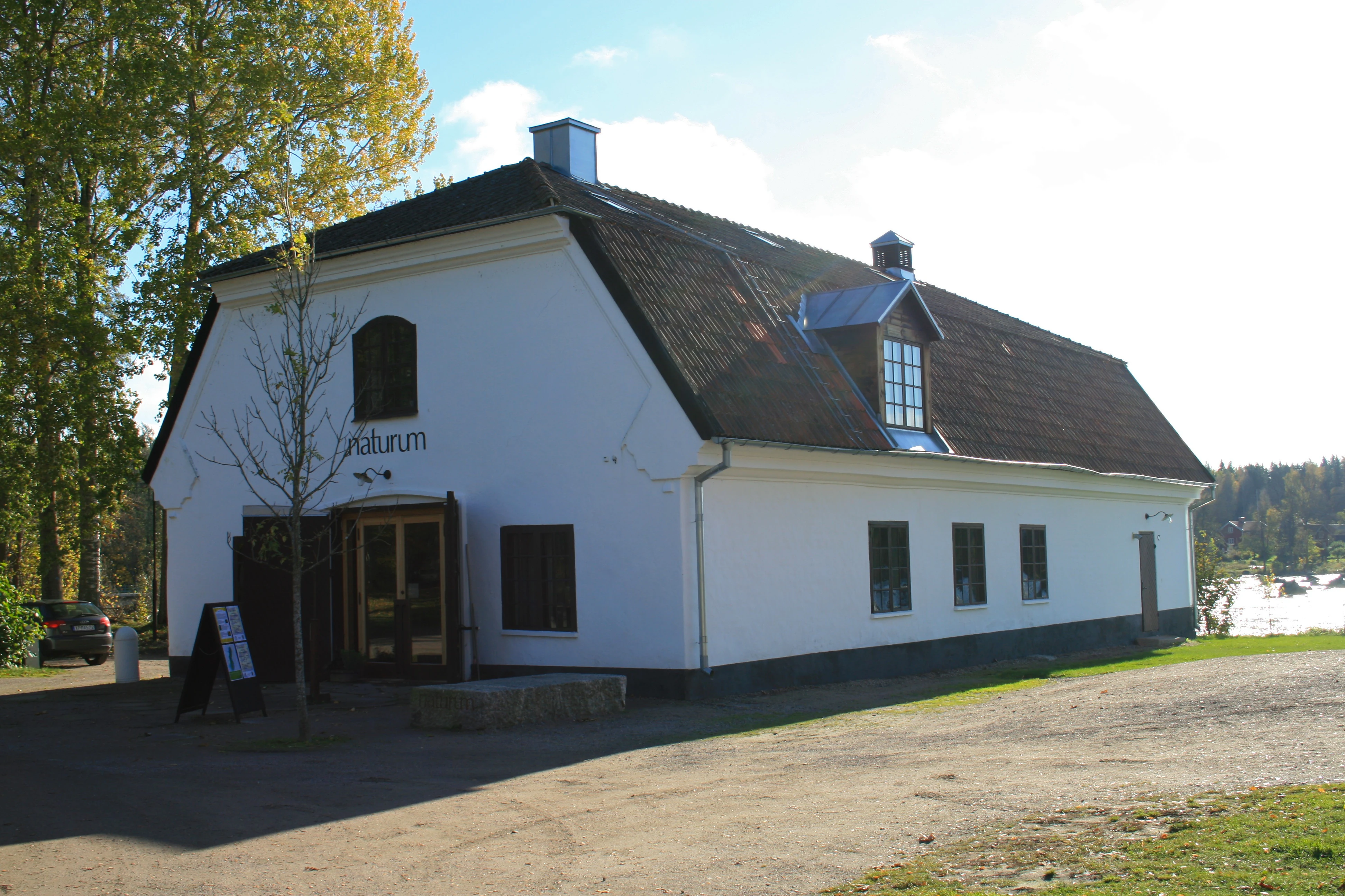 White building with brown roof, 'Naturum' text on facade, surrounded by trees and open ground with water body visible in background