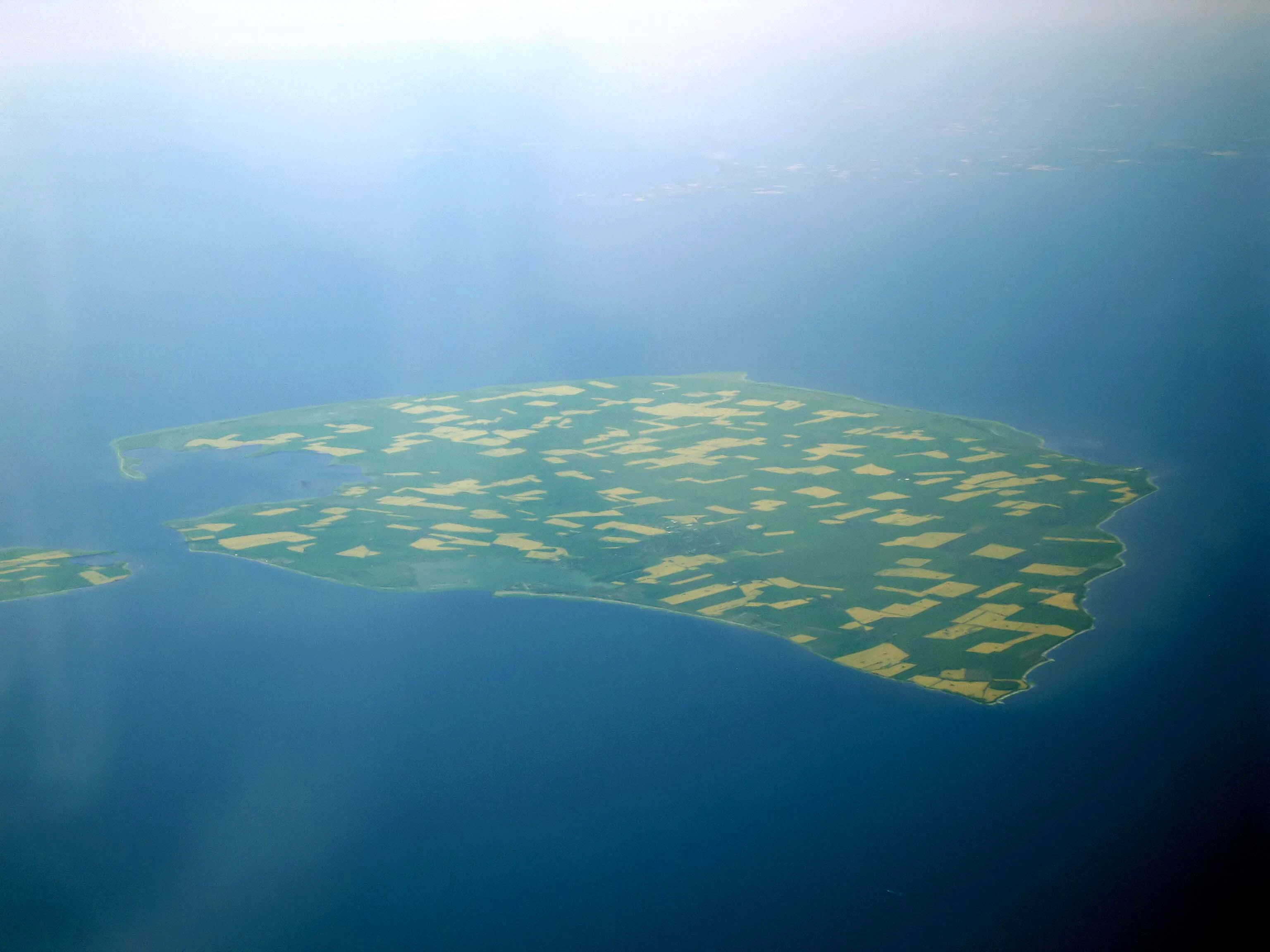 An aerial photograph of the island of Fehmarn, showing a flat, green landscape divided into small agricultural fields, entirely surrounded by the dark blue waters of the Baltic Sea.