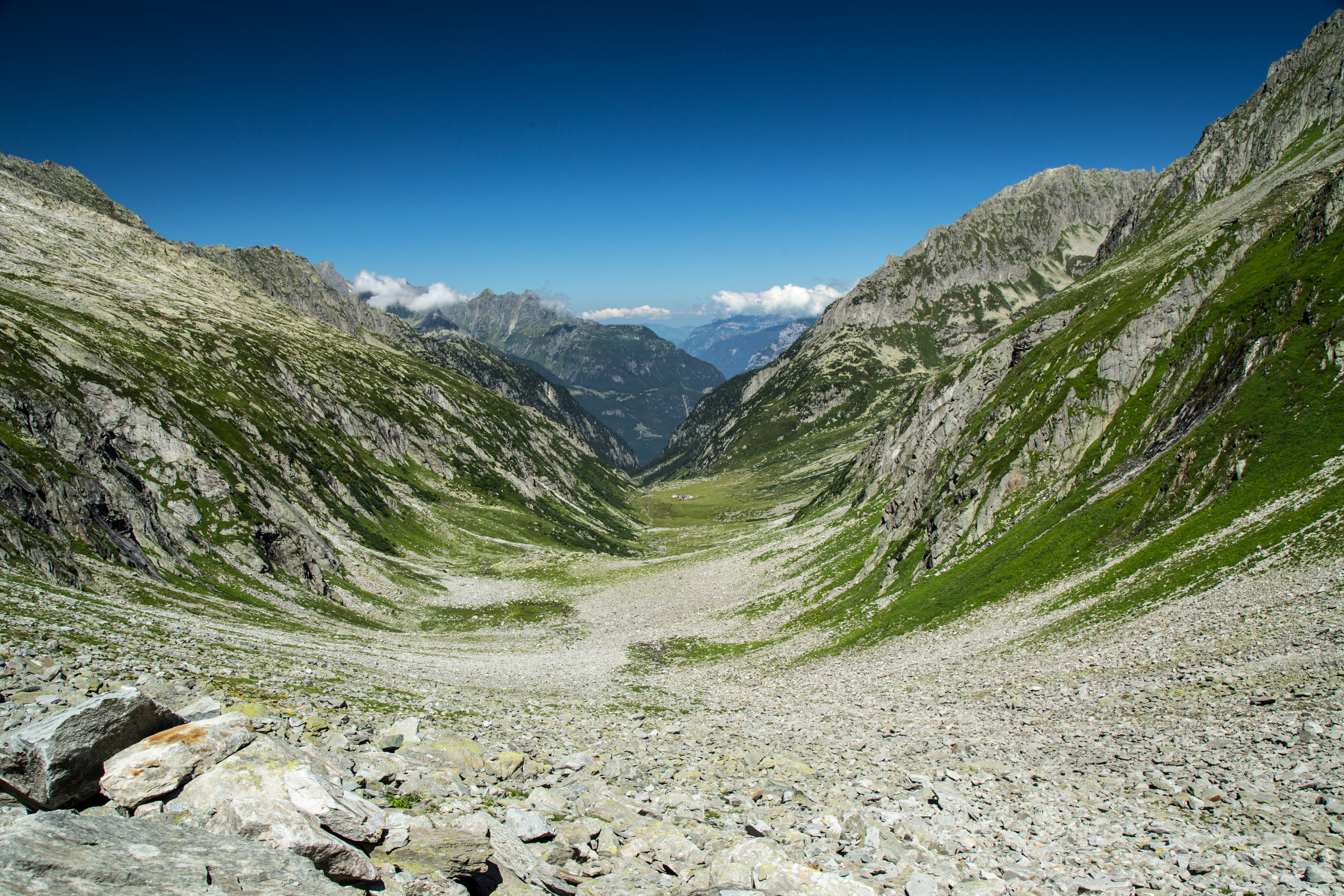 An eye-level wide shot of the glaciated Fellital valley in the Swiss Alps, showing a rocky mountain pass descent leading into a verdant valley floor flanked by steep, partially green rocky slopes under a clear blue sky.
