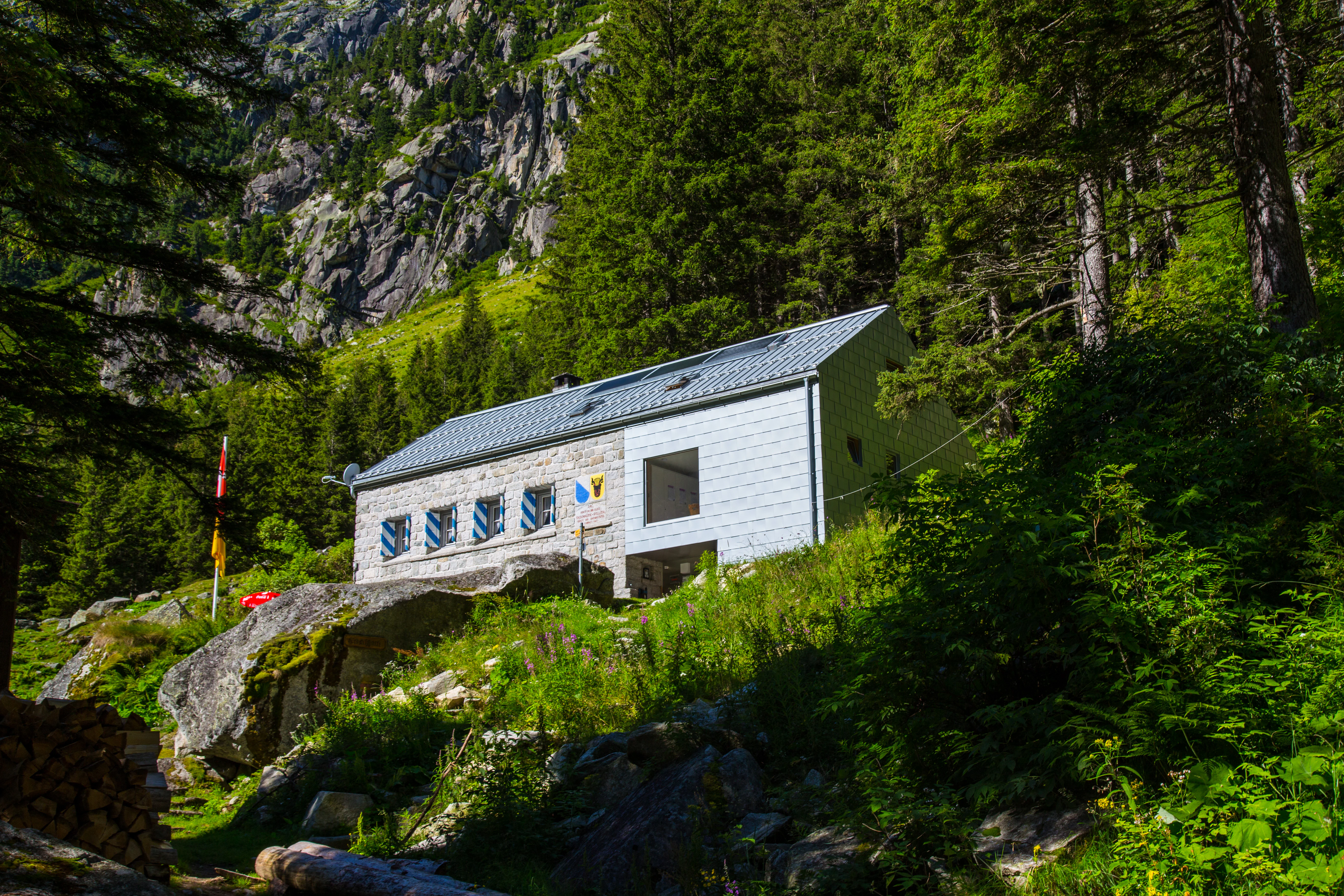 A two-story mountain hut with a stone and gray facade sits on a grassy, rock-strewn slope surrounded by a dense coniferous forest and steep mountain cliffs.