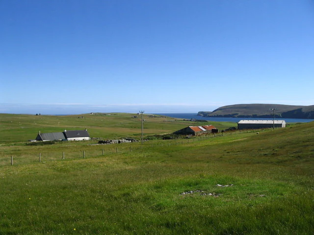A wide view of a green, grassy landscape on Fetlar, Scotland, featuring scattered stone buildings, farm structures, rolling hills, and a glimpse of the ocean in the distance under a clear blue sky.