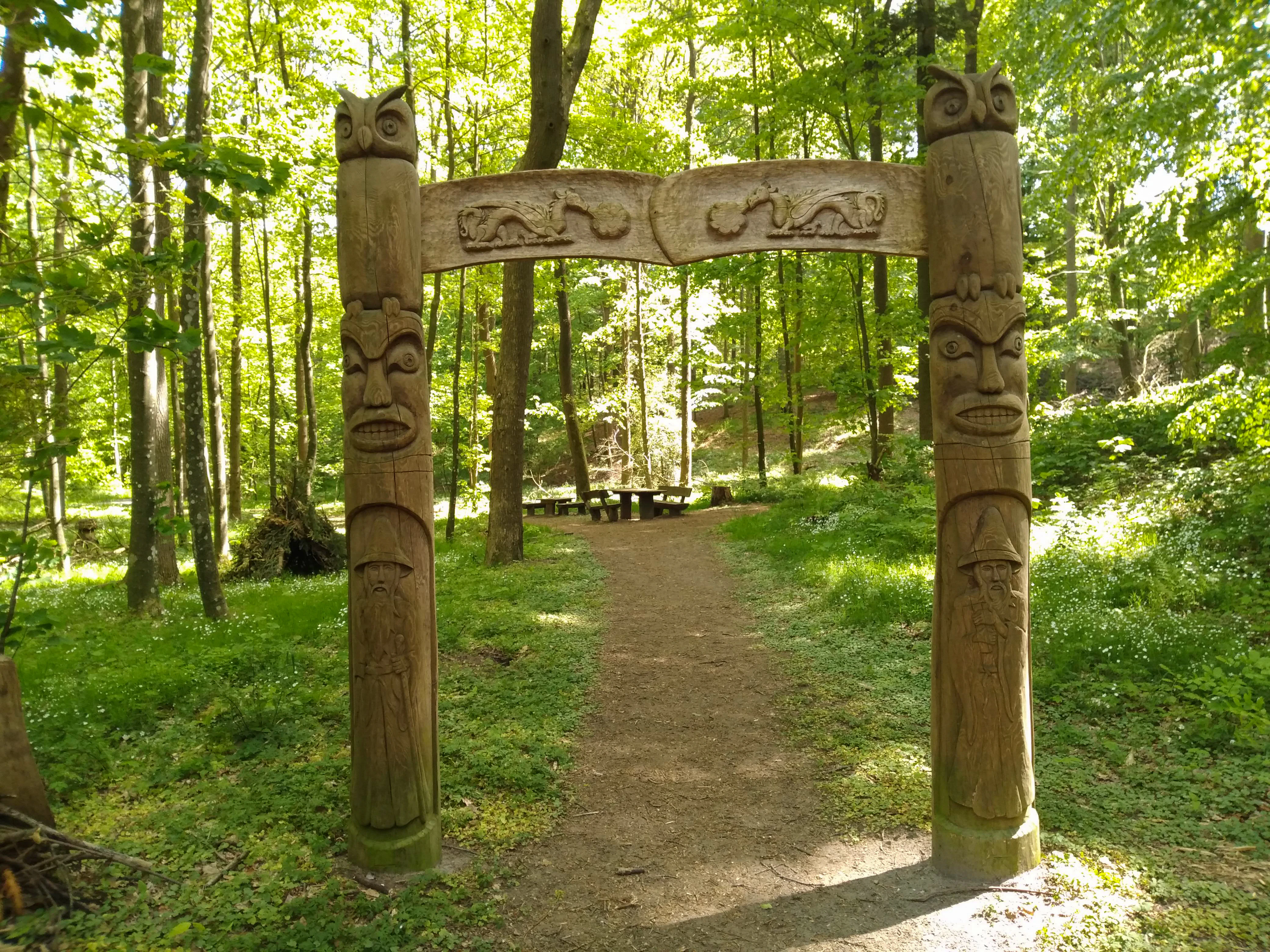 A decorative wooden archway featuring carved faces and animal figures marks the entrance to a forest trail, with a picnic table visible in the distance.