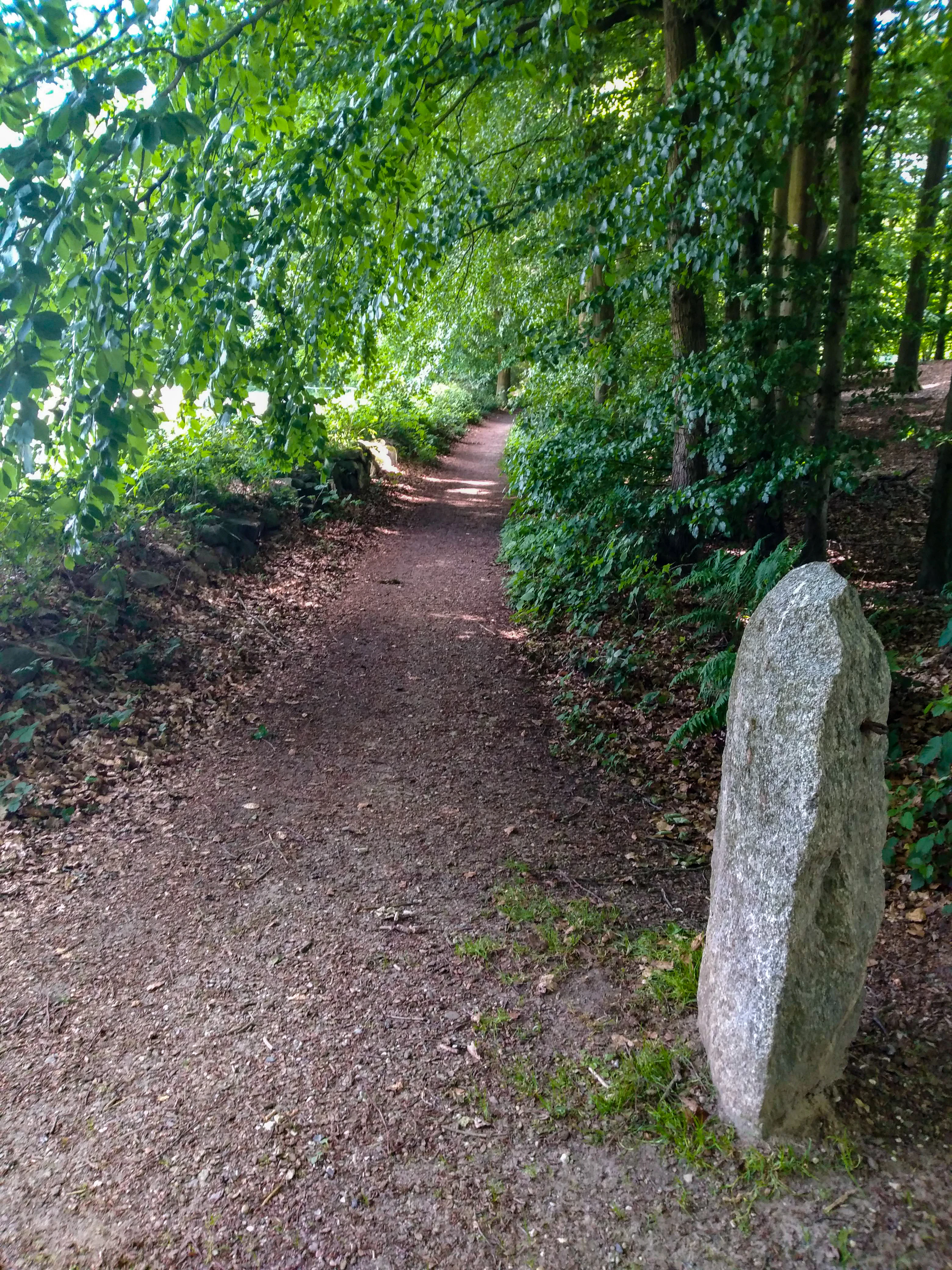 A narrow dirt hiking path winding through a lush green forest, with a prominent weathered stone marker standing on the right side of the trail.