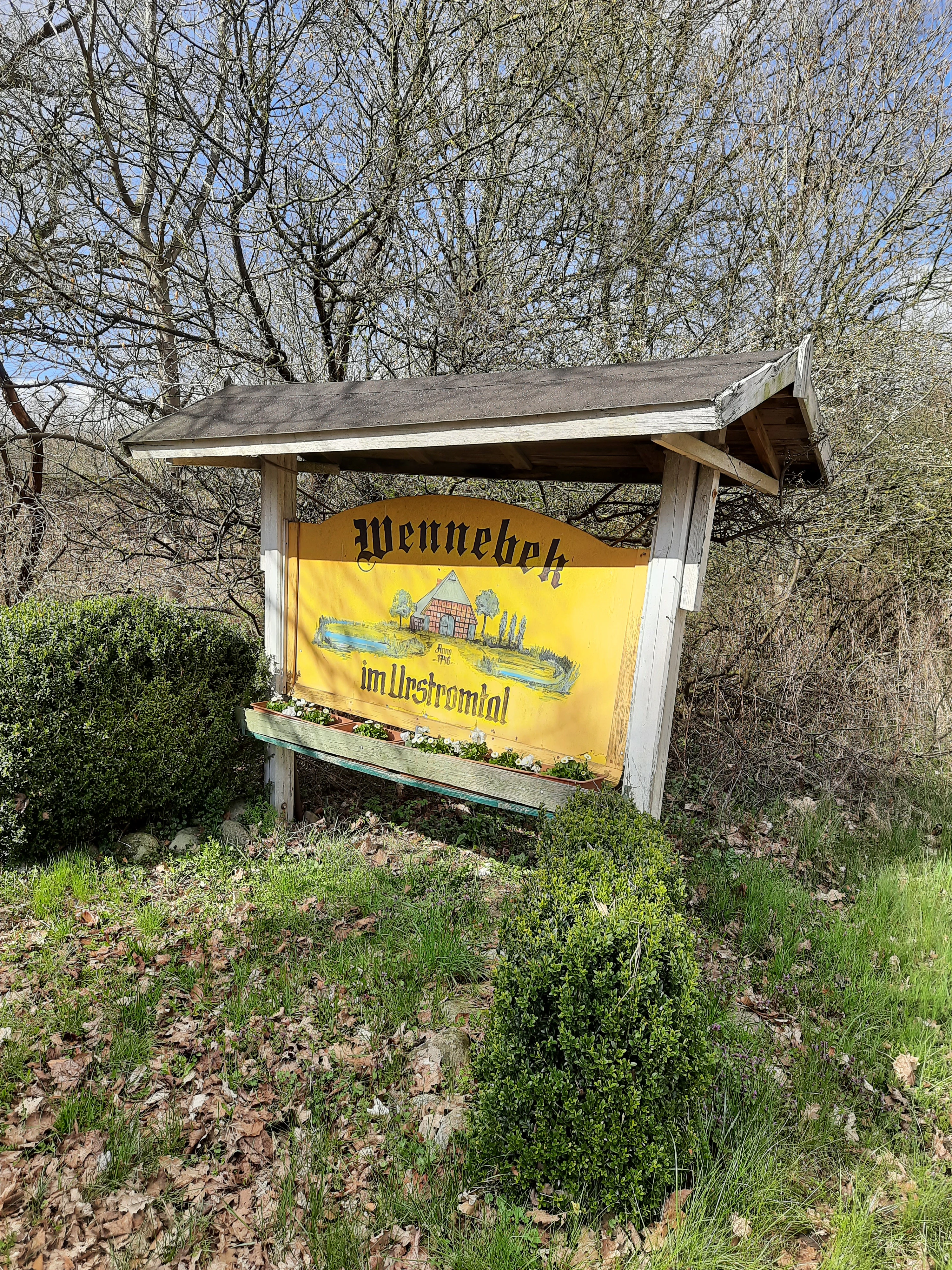 A weathered yellow information sign with a wooden roof stands in a field; it features an illustration of a traditional house and the text Wennebek im Urstromtal.