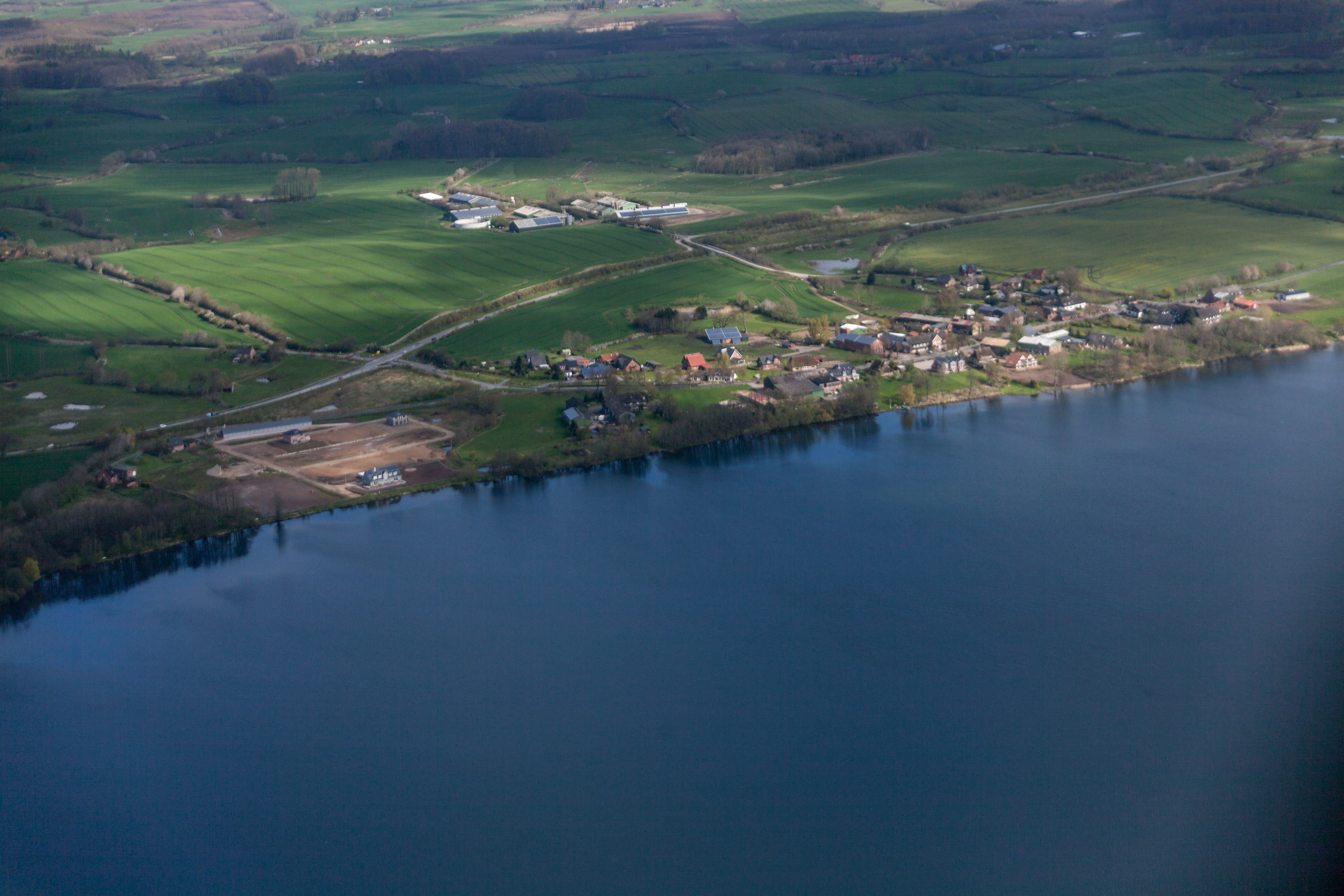 An aerial view showing a large dark blue lake bordered by a small village, green fields, and rural roads.