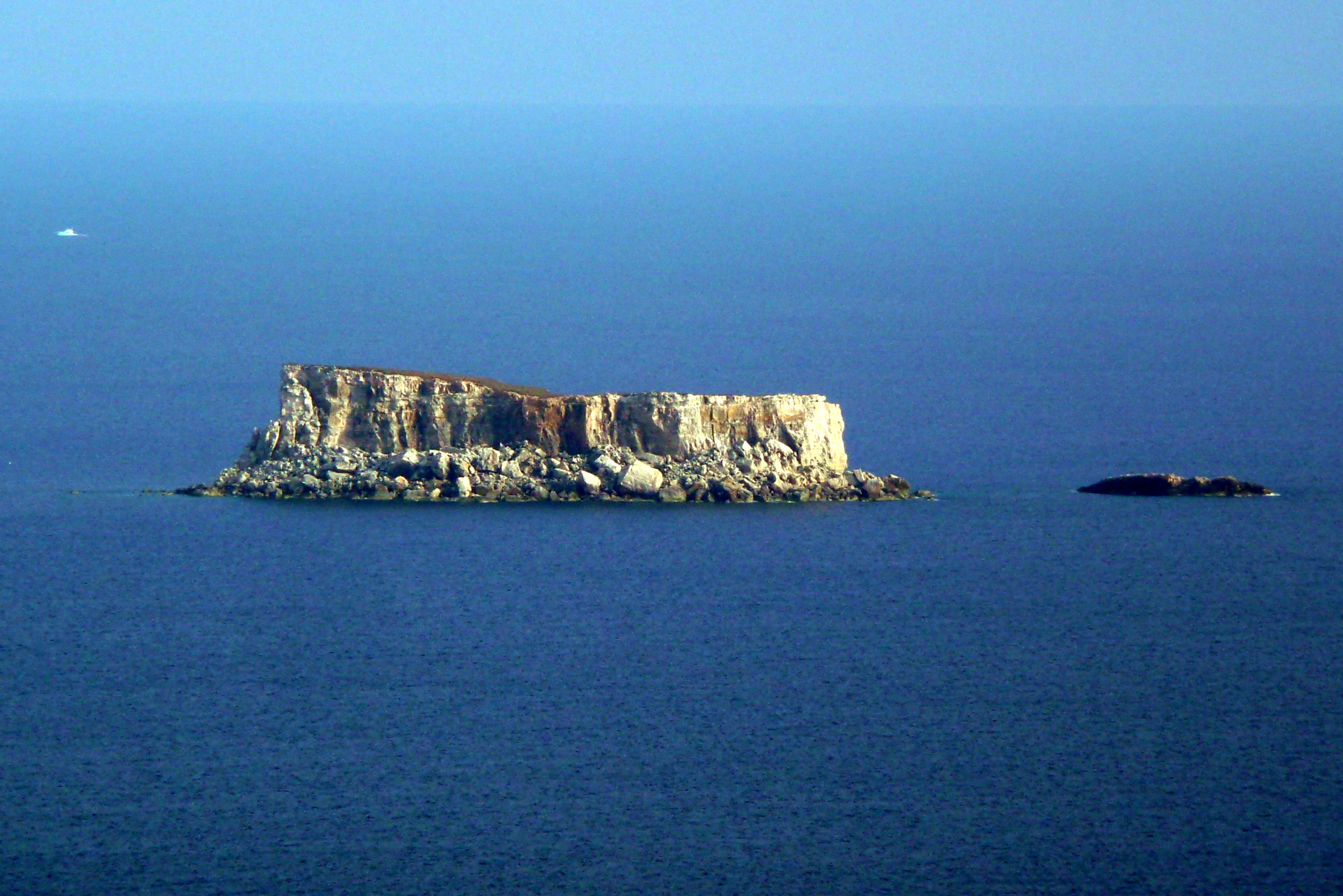 A distant, eye level view of the small, flat topped limestone islet of Filfla, surrounded by deep blue water, with a smaller rock formation visible to its right.