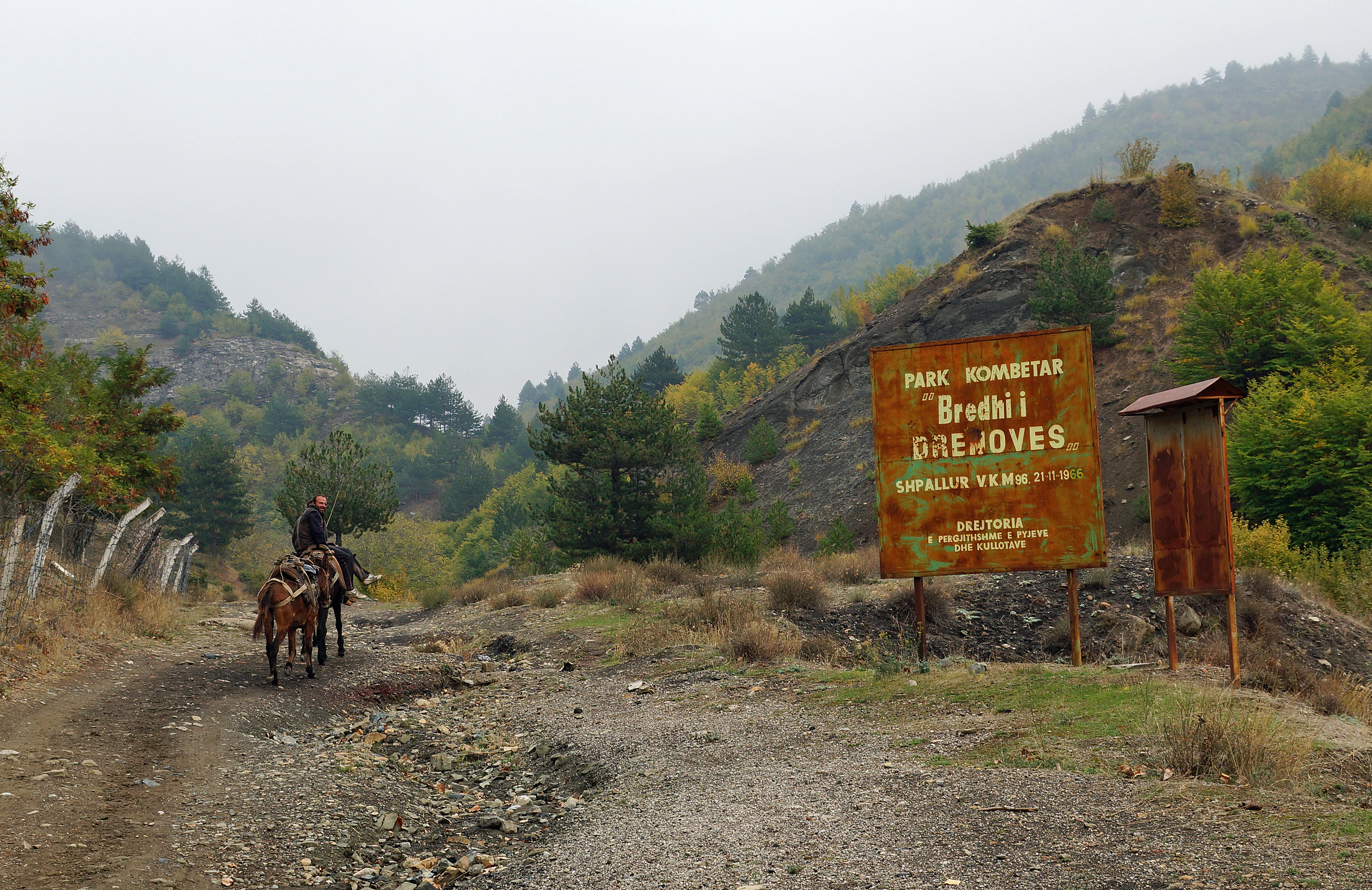 A weathered, rusted metal sign marking the Fir of Drenovë national park stands on a dirt road in a mountainous, forested landscape, with a person on horseback approaching from the left.