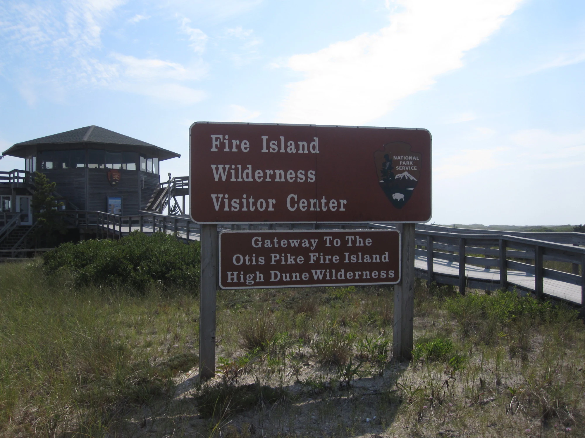 A brown National Park Service sign identifying the Fire Island Wilderness Visitor Center and the entrance to the Otis Pike Fire Island High Dune Wilderness, with the wooden visitor center building in the background.