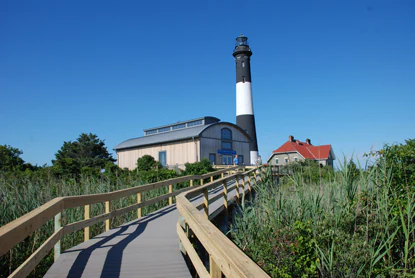 A view of the Fire Island Lighthouse with a black and white tower, connected to a visitor building, accessed by a wooden boardwalk winding through tall green grasses under a clear blue sky.