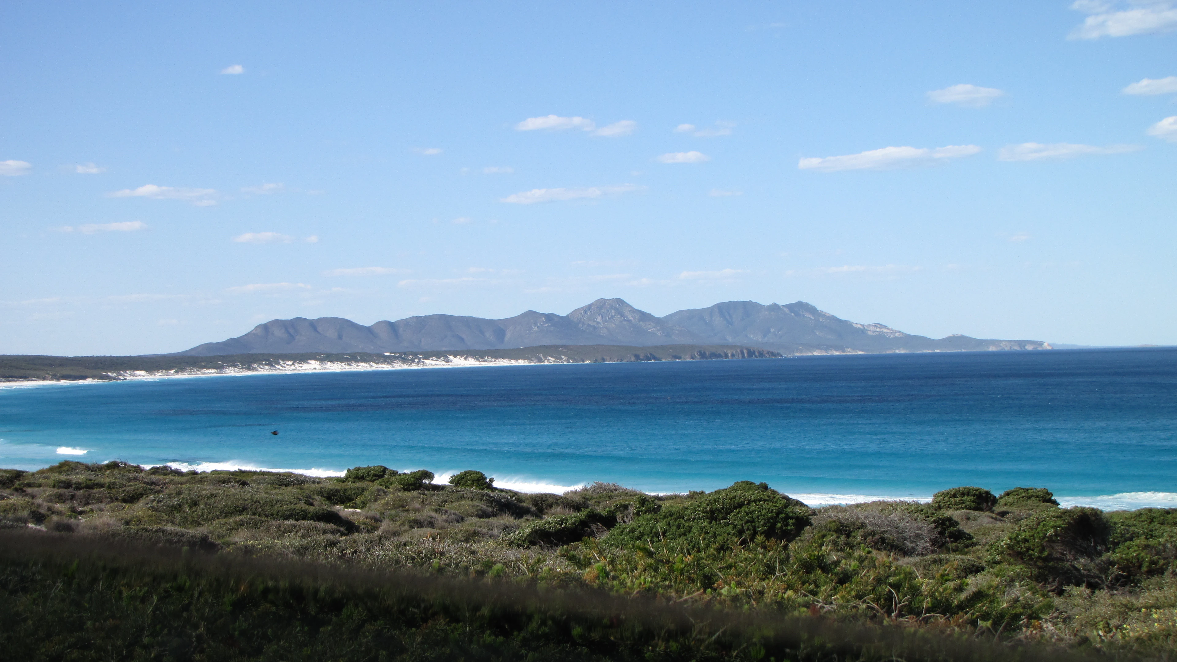Coastal landscape with blue ocean, distant mountains, and green vegetation under a clear sky