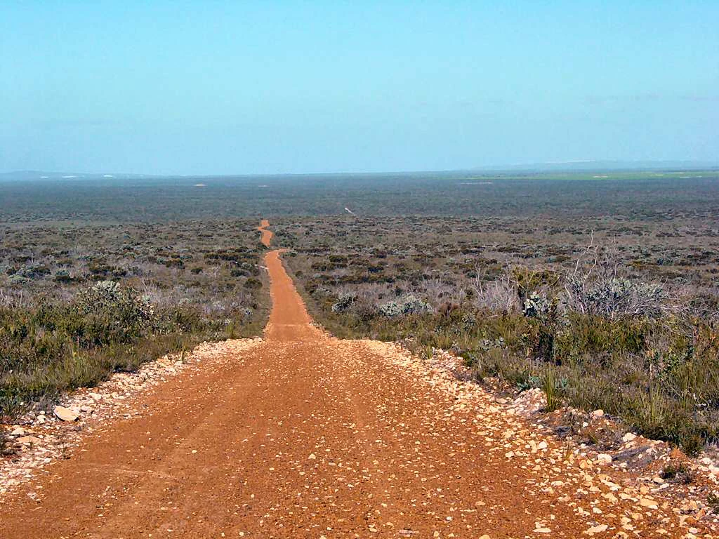 A dirt road running through a vast, open landscape with sparse vegetation under a clear blue sky