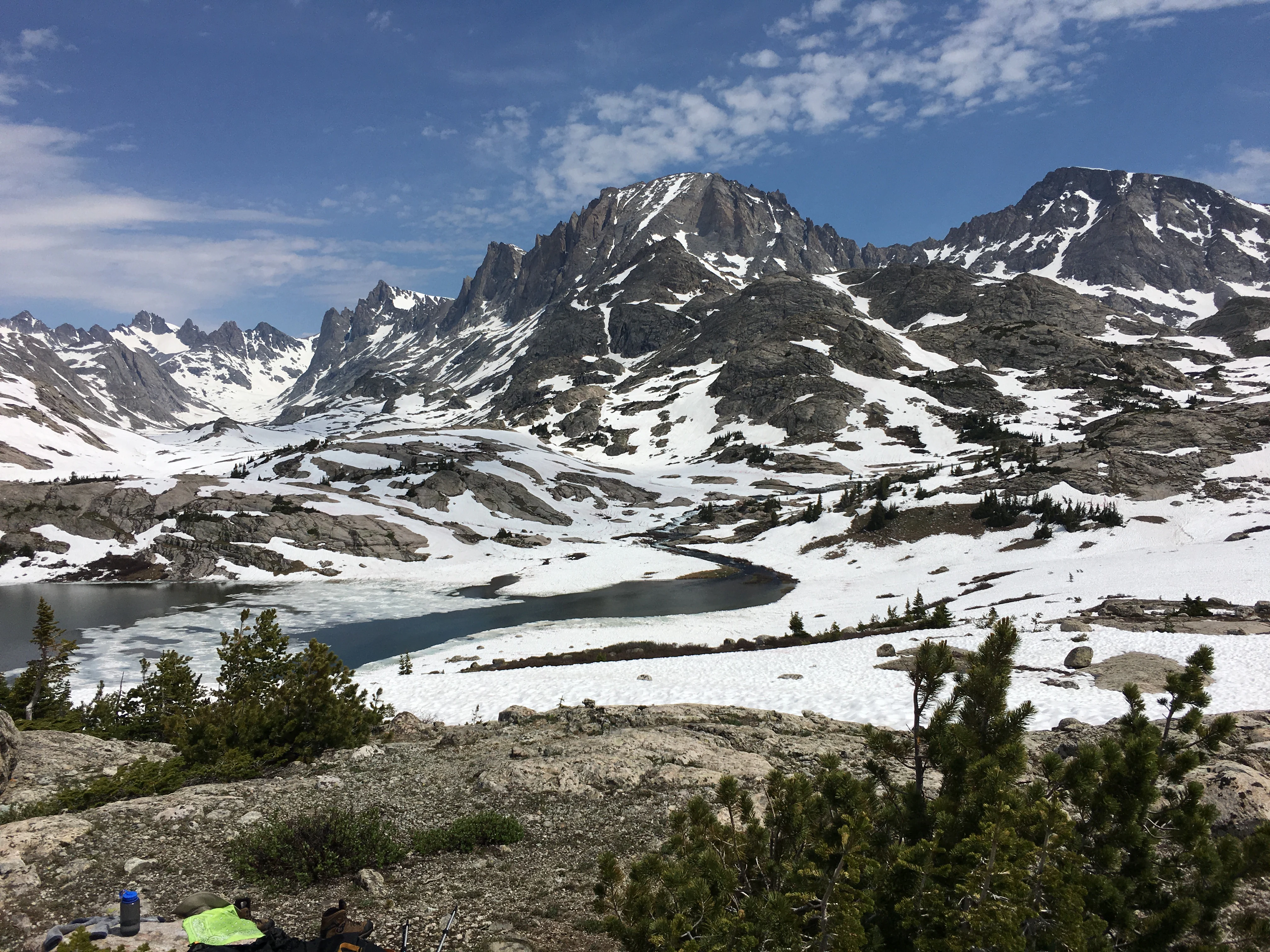 A vast, rugged mountain landscape with jagged, snow-dusted granite peaks reflecting in a partially frozen alpine lake under a blue sky with light clouds.