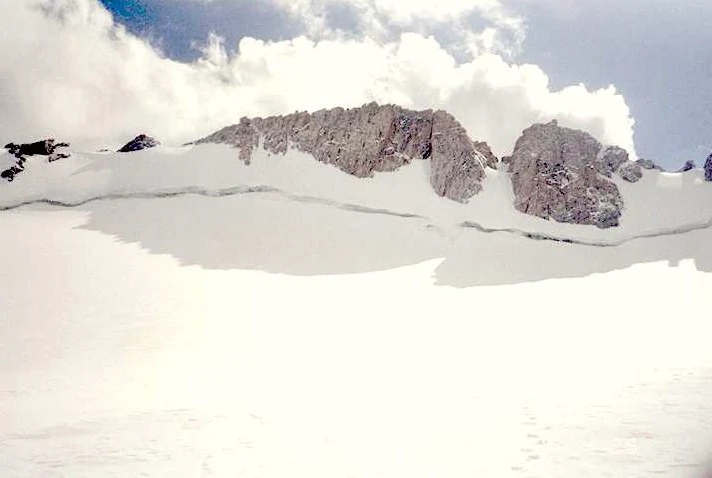 A wide shot of a high altitude glacier covered in snow with rocky, jagged mountain peaks rising above the snow line against a cloudy sky.